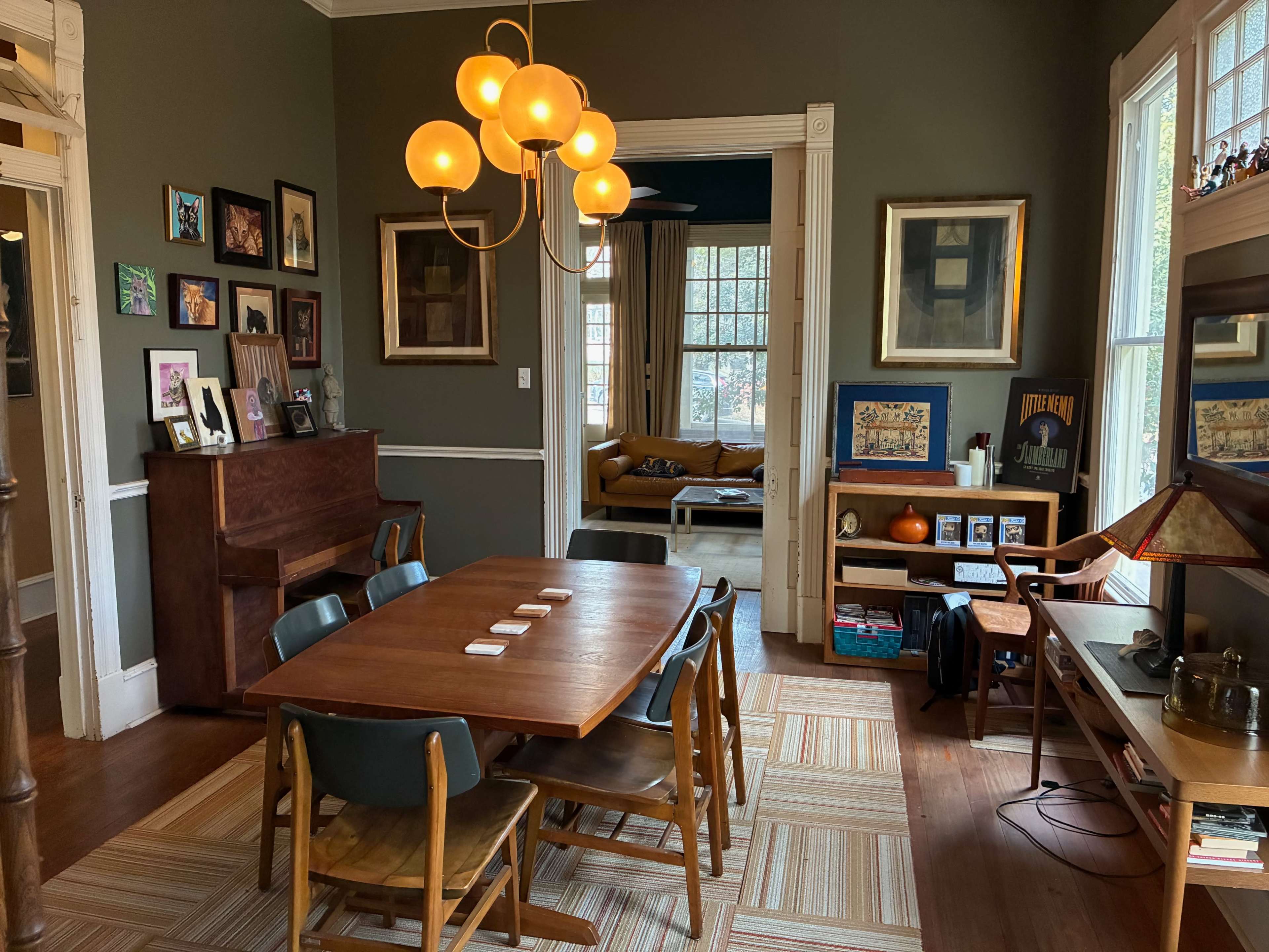 The image shows a dining area with a wooden table surrounded by chairs, a piano against the wall, and a cozy living room visible through a doorway.