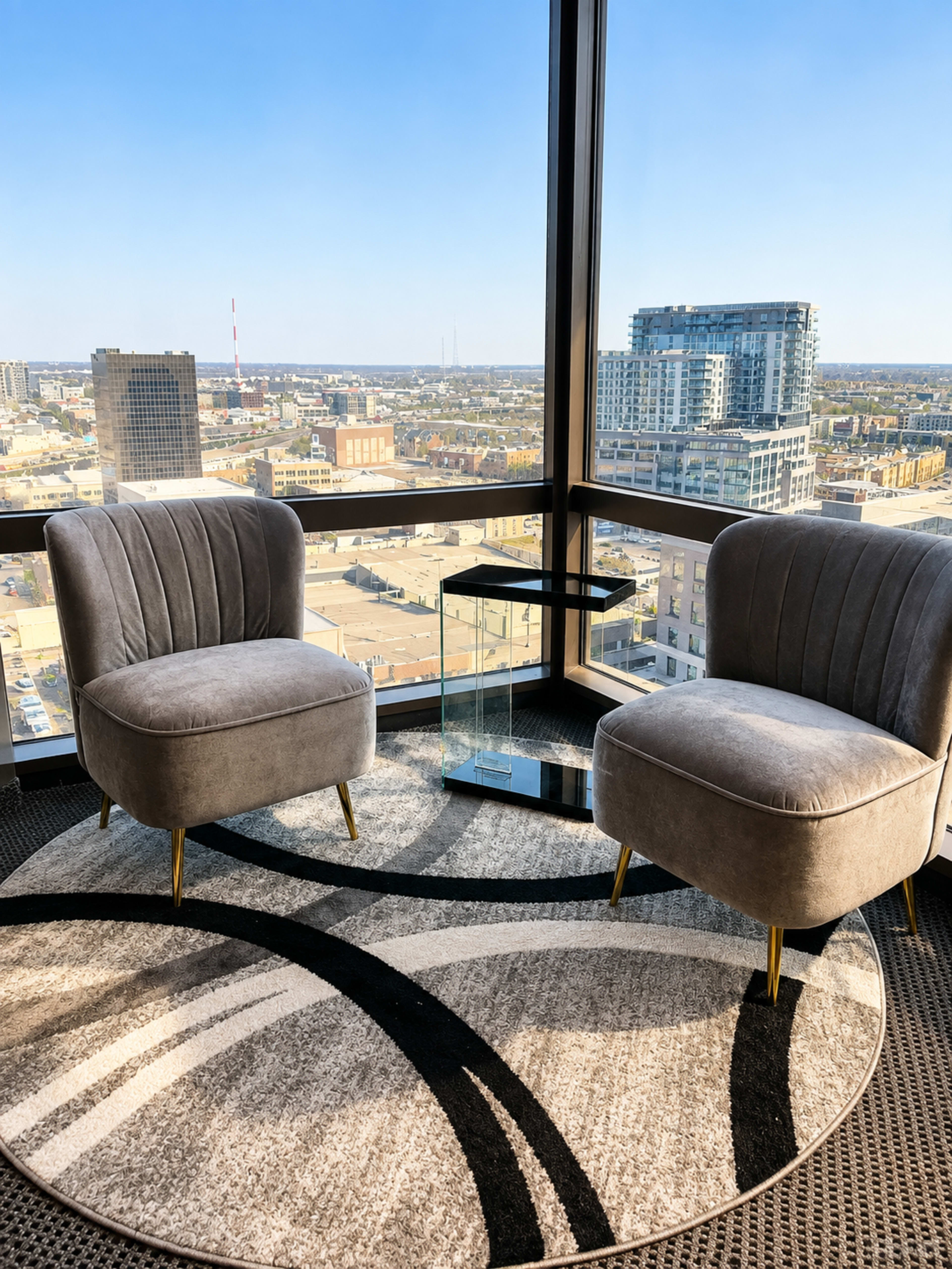 Two gray upholstered chairs are positioned beside a glass side table on a circular geometric rug, overlooking a city skyline through large windows.