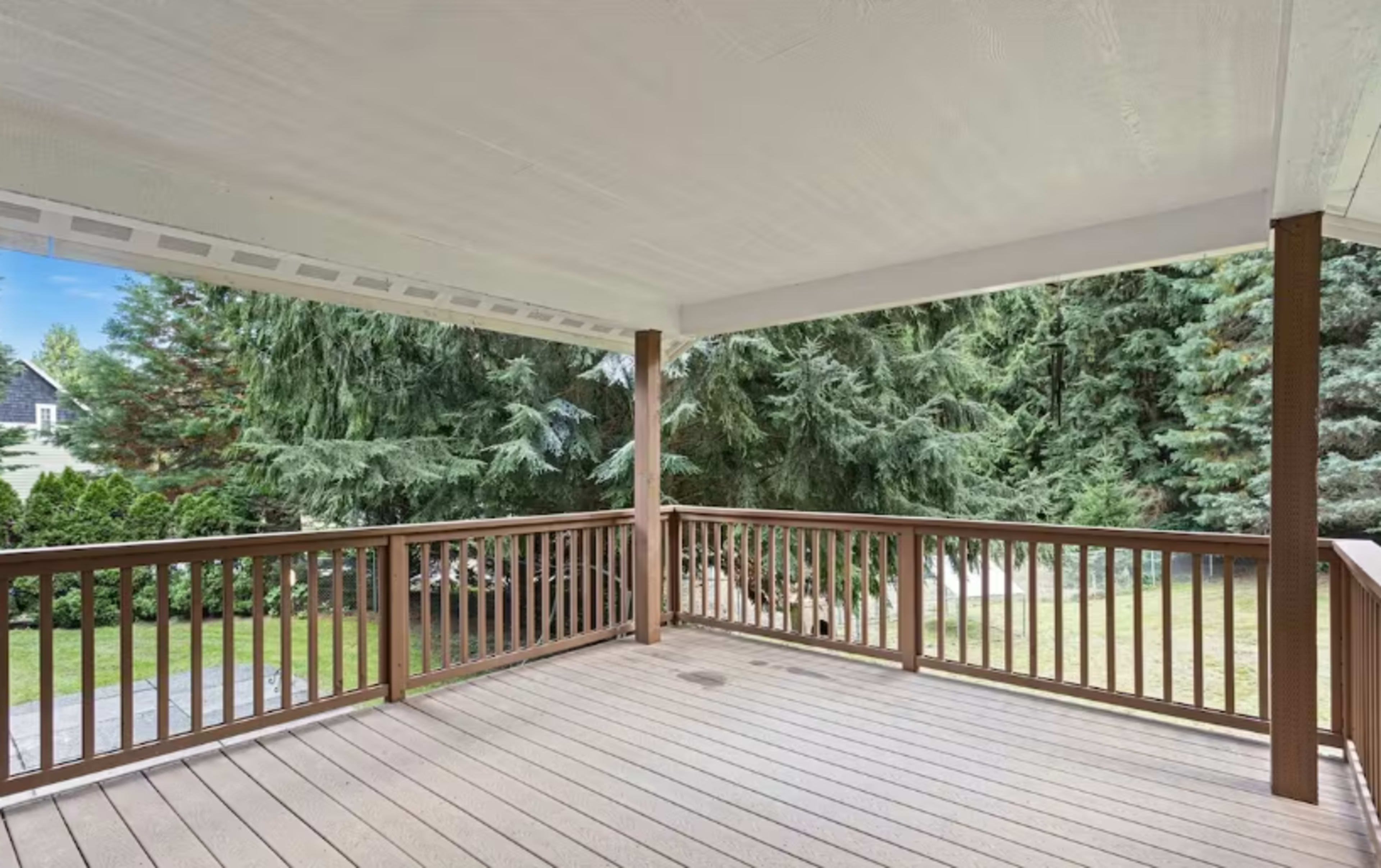 The image shows a wooden deck with a covered roof, surrounded by tall green trees and a grassy area.