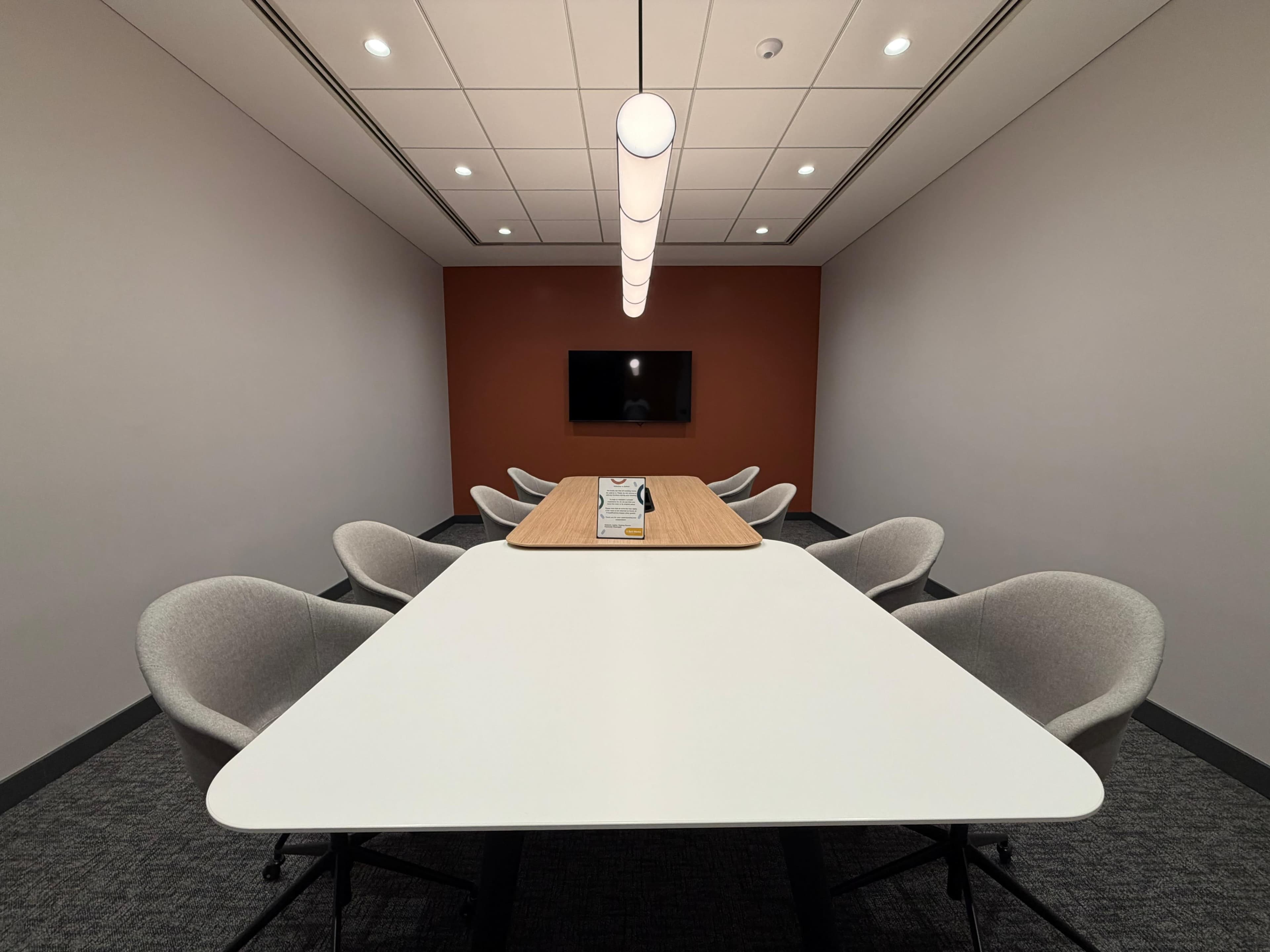 A modern conference room features a long table surrounded by ergonomic chairs, with a large screen mounted on the wall and a pendant light overhead.