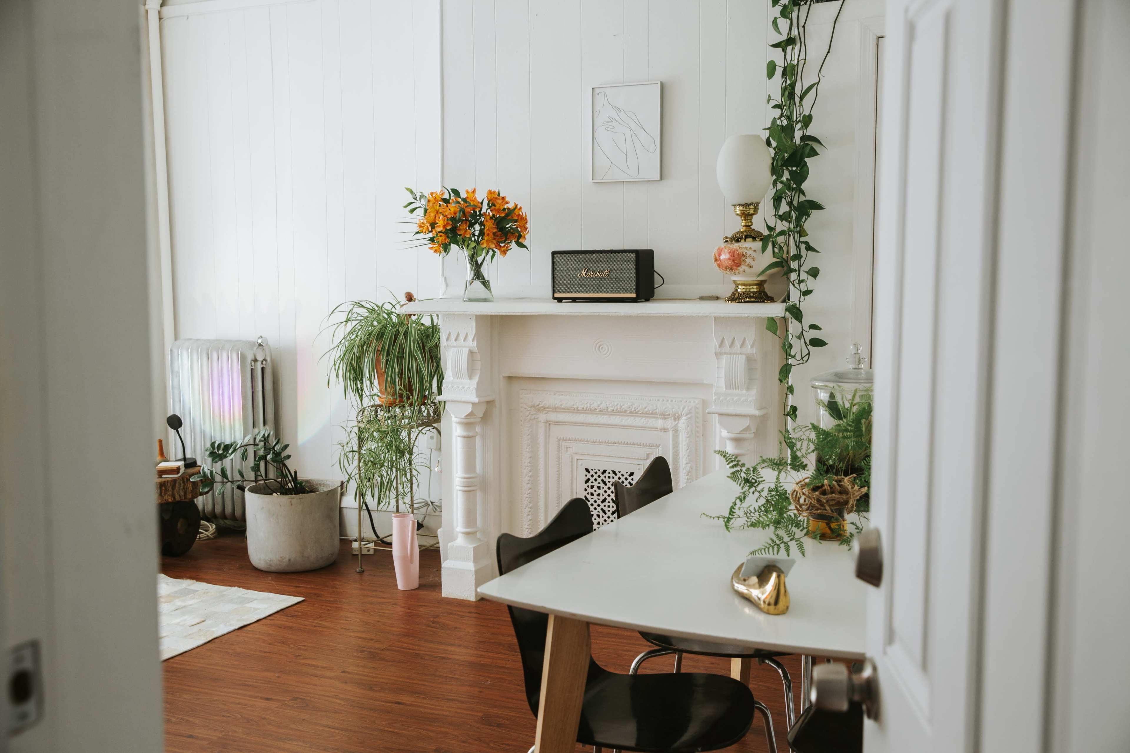A bright, minimalist living space features a white mantel with decorative plants and a vintage radio, adjacent to a sleek dining table and a doorway leading to another room.
