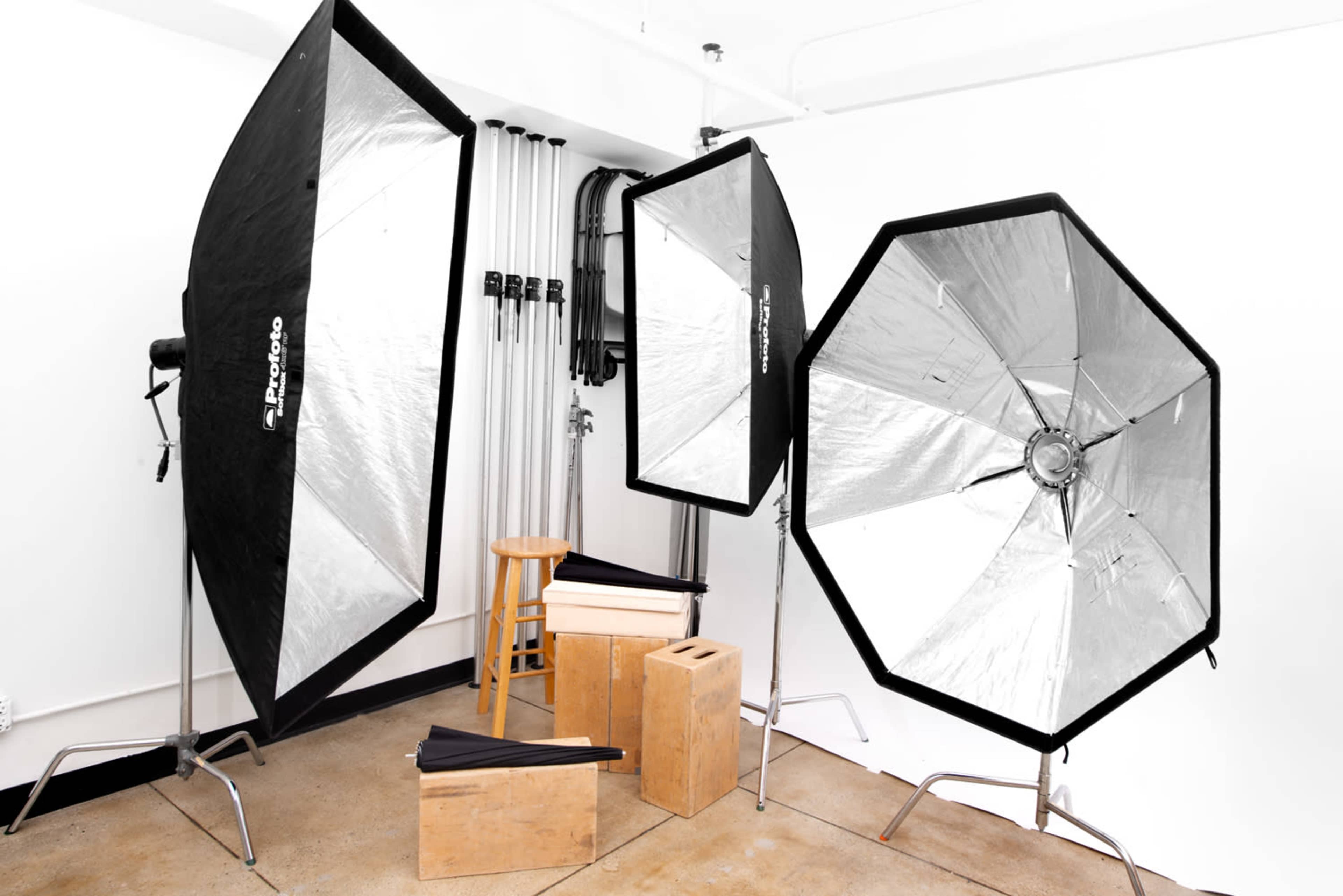 A photography setup with three light modifiers, a stool, and various wooden boxes against a white wall.