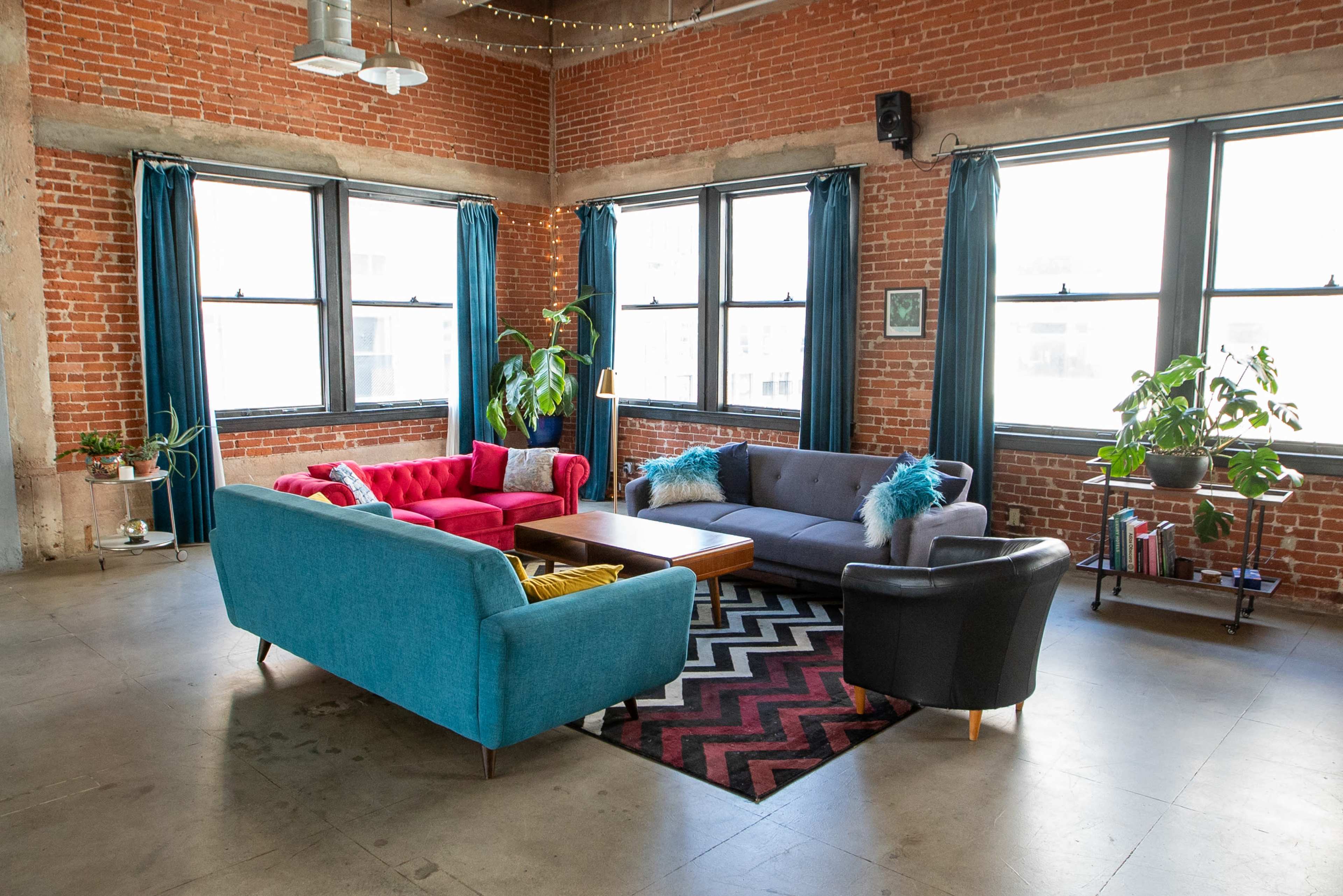 A modern living room with a blue and pink couch, a black chair, a wooden coffee table, and several potted plants, all set against a brick wall with large windows.