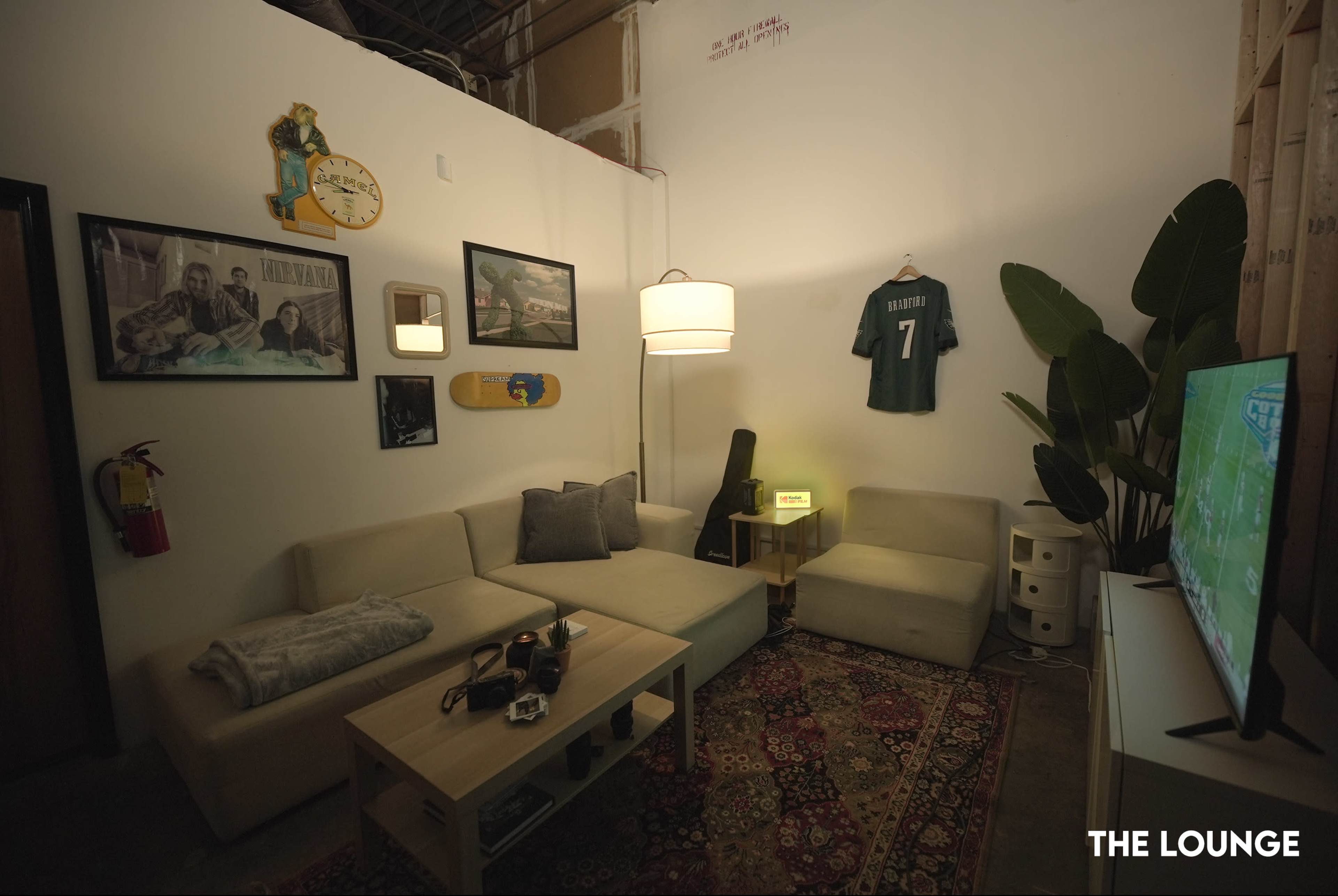 The image shows a cozy lounge area with a light-colored sectional sofa, a rug, a coffee table, and various wall decorations, including a clock and framed pictures.
