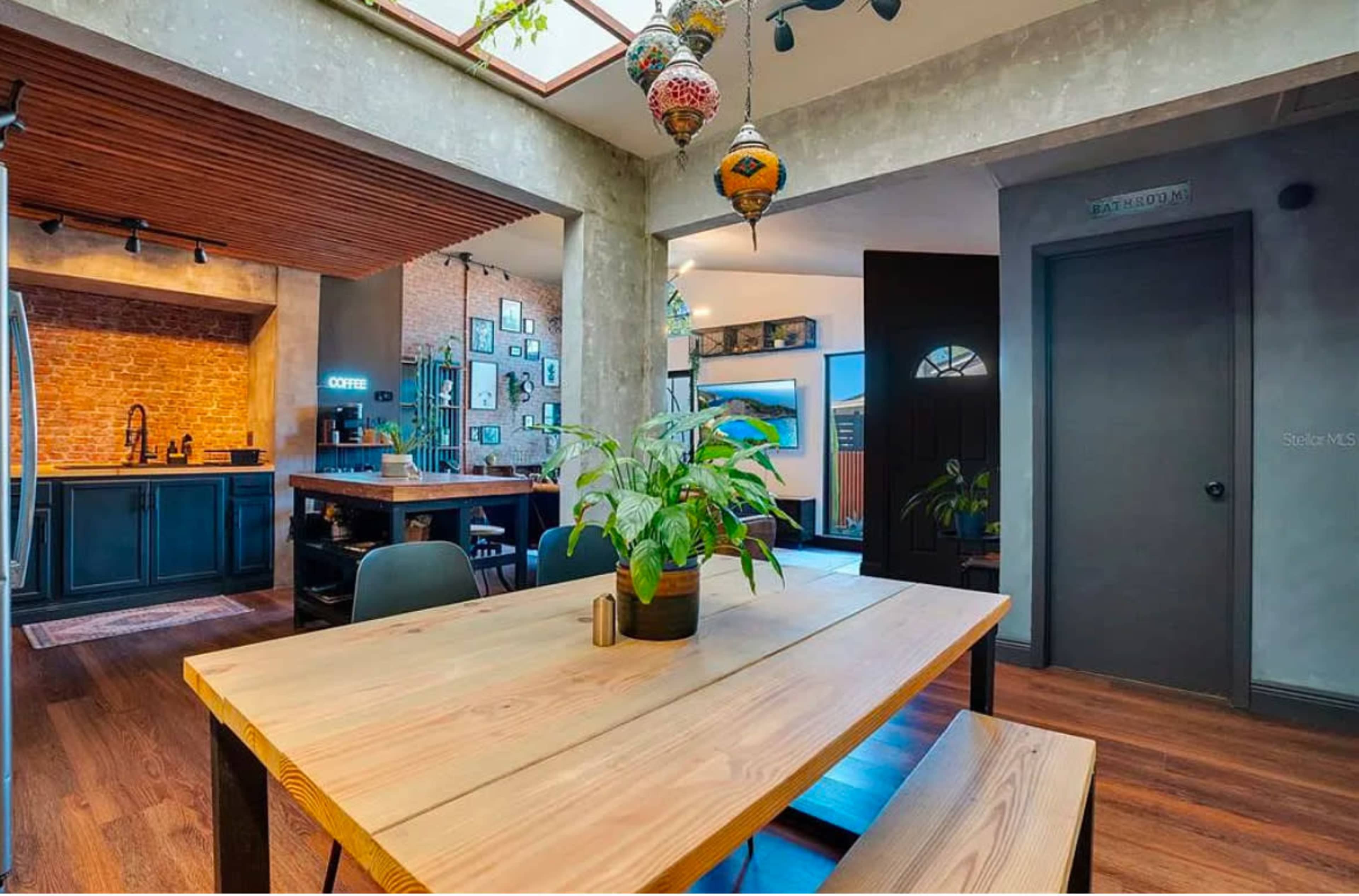 The image shows a modern, minimalistic kitchen and dining area featuring a wooden table surrounded by chairs, with exposed brick walls and hanging decorations.