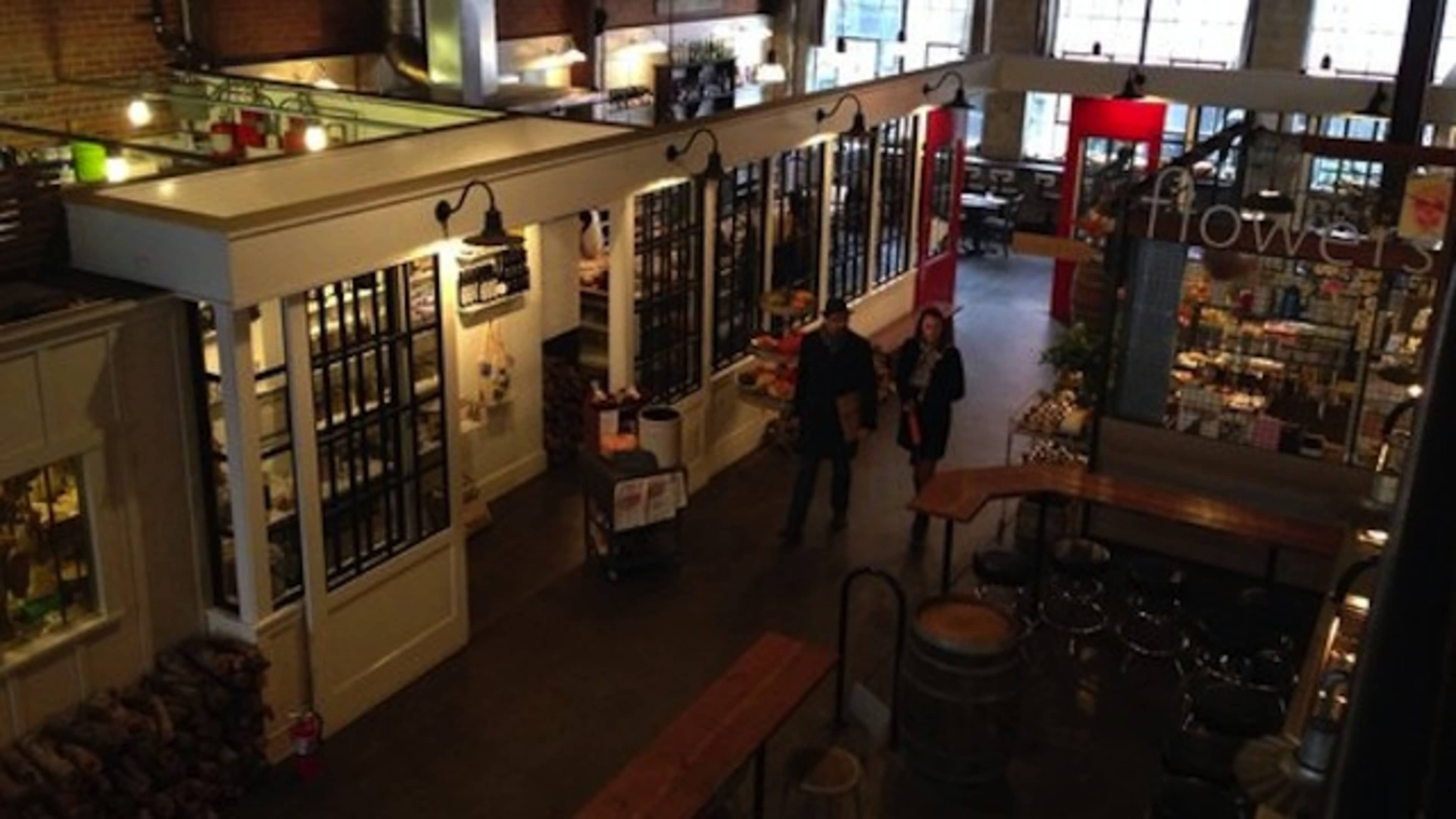 The image shows a bustling indoor market with wooden tables, various stalls, and large windows letting in natural light.