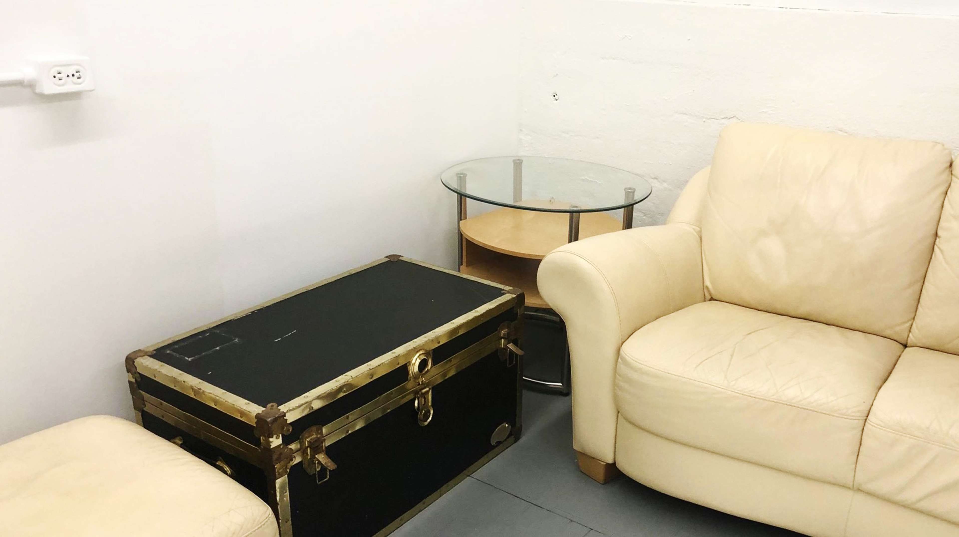 The image shows a small room with a light-colored leather couch, a glass-top side table, and a black trunk.