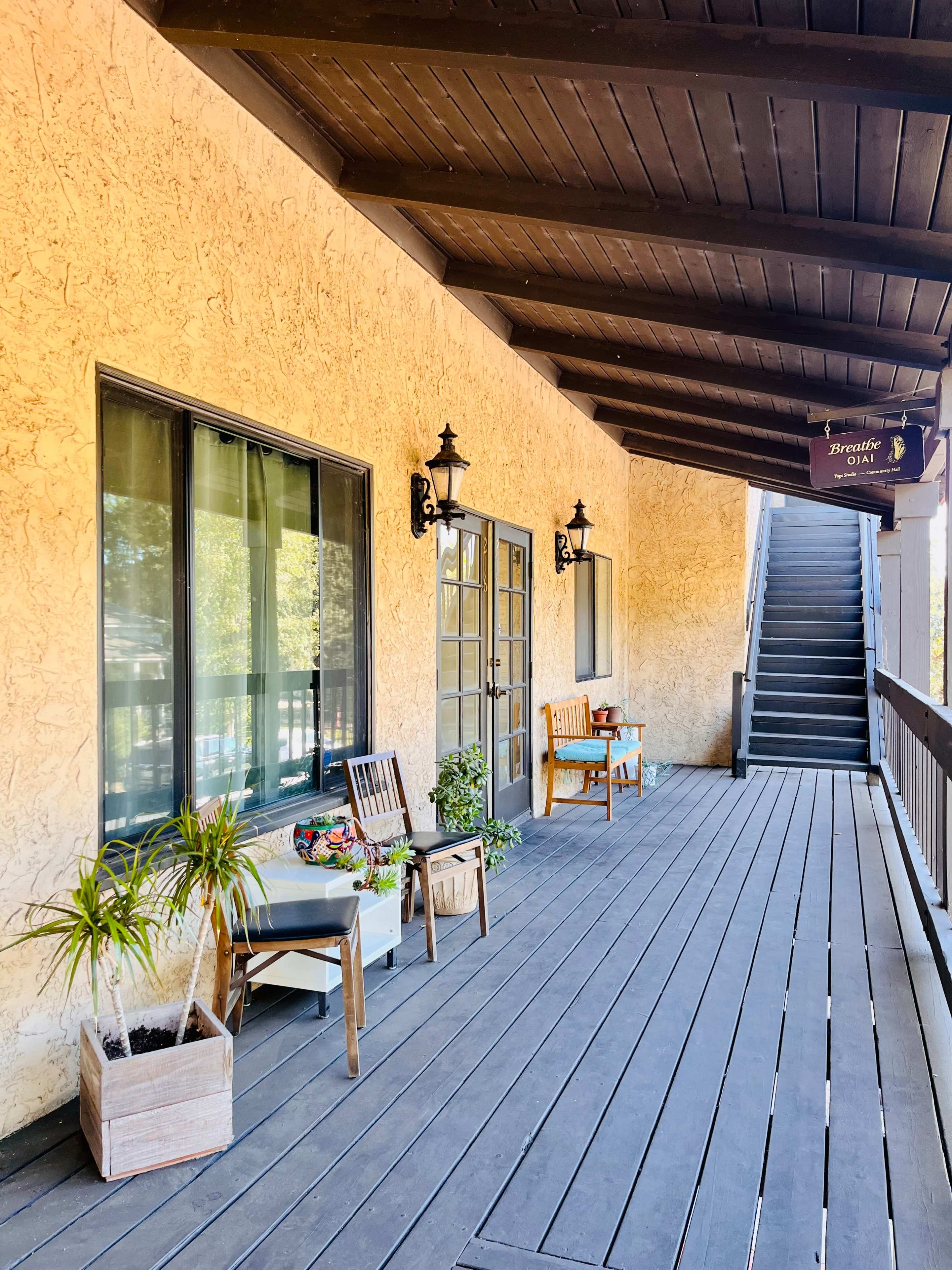 The image shows a covered wooden walkway with two chairs and potted plants beside a pair of glass doors, leading to a staircase.