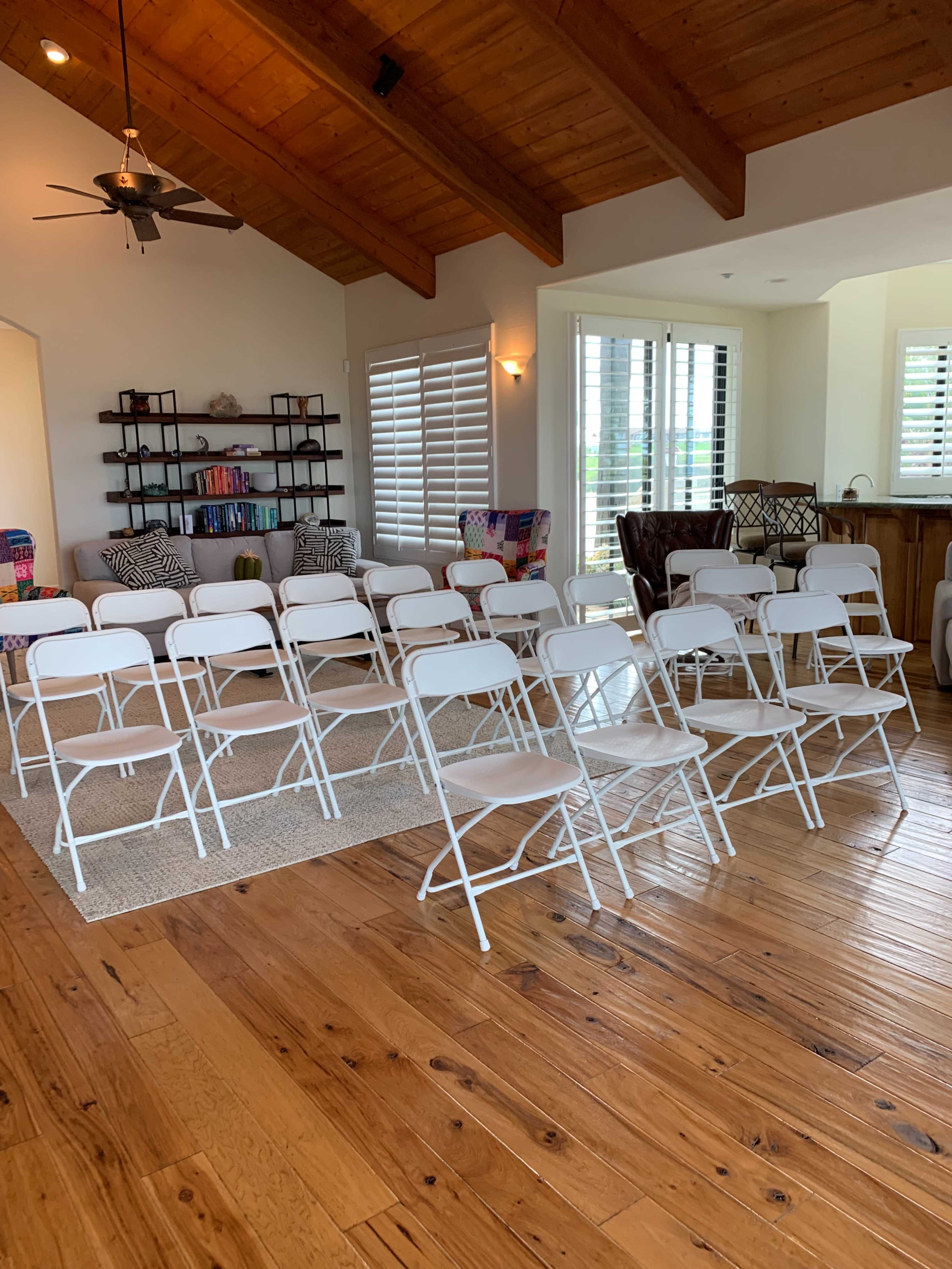 A room with wooden floors and a high ceiling has several rows of white folding chairs arranged in front of a bookshelf and a seating area.