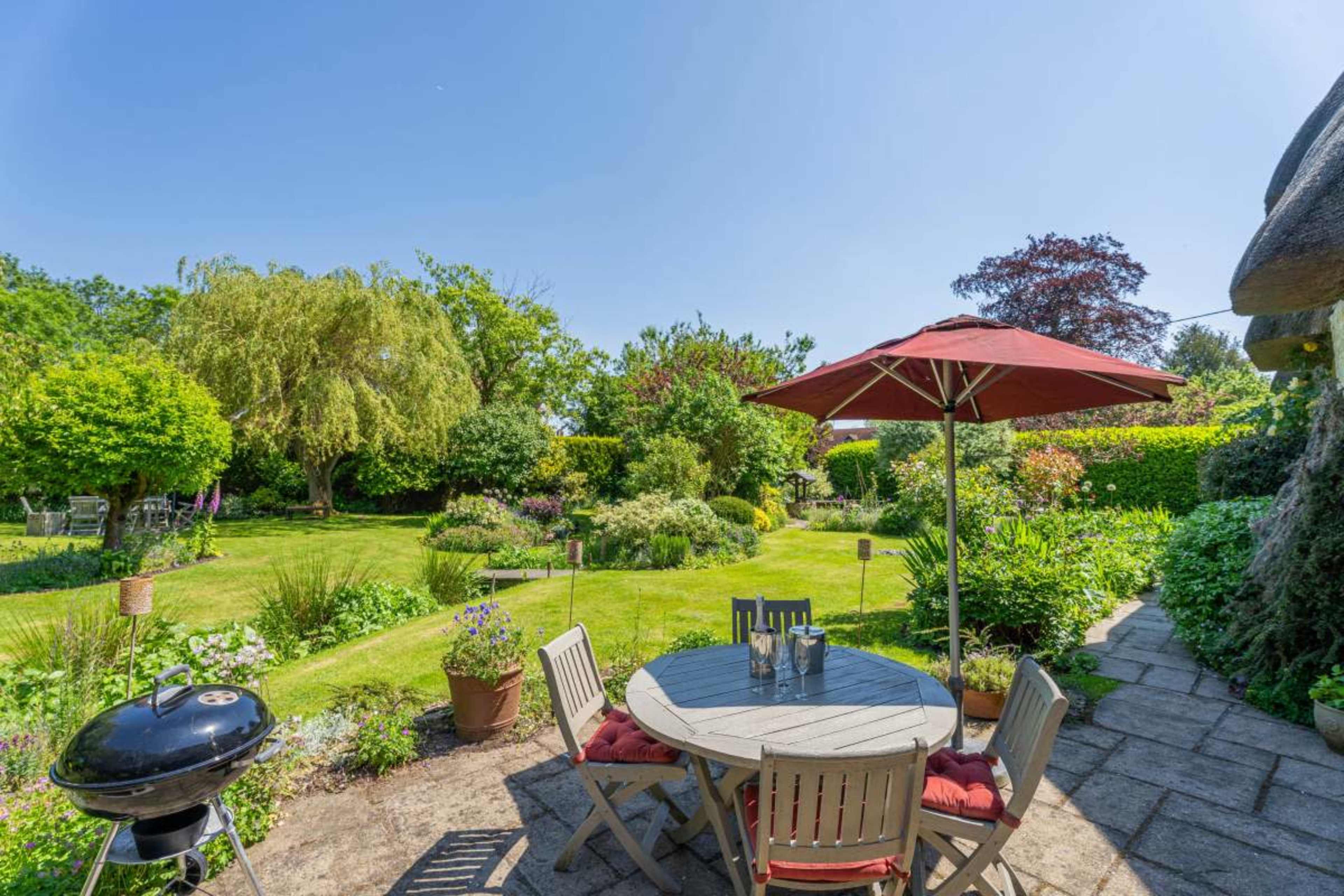 A patio with a round table and chairs is set up under a red umbrella, overlooking a lush garden with various plants and a charcoal grill nearby.