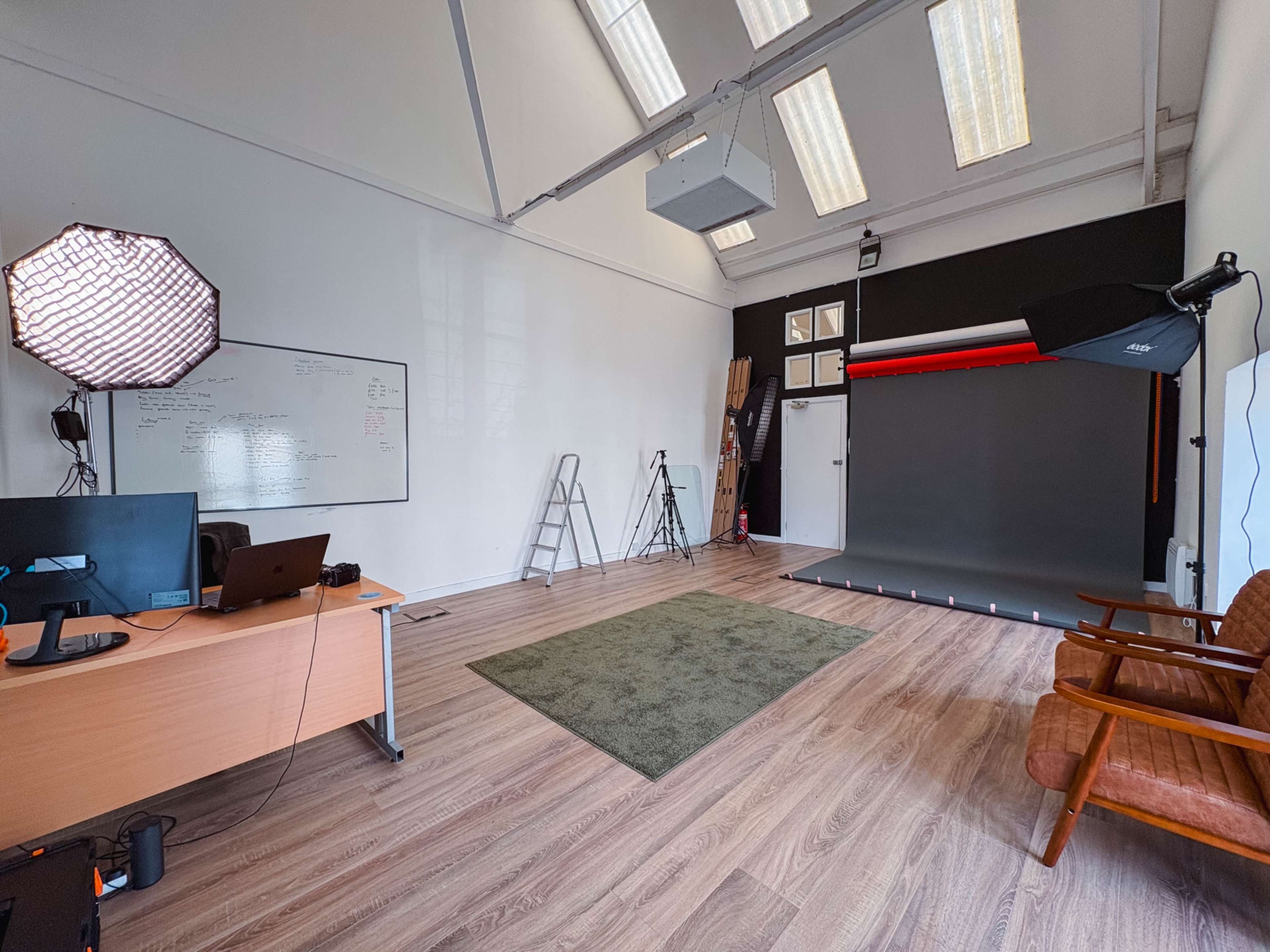 A spacious photography studio features a green area rug, a large backdrop, a desk with a computer, and seating.