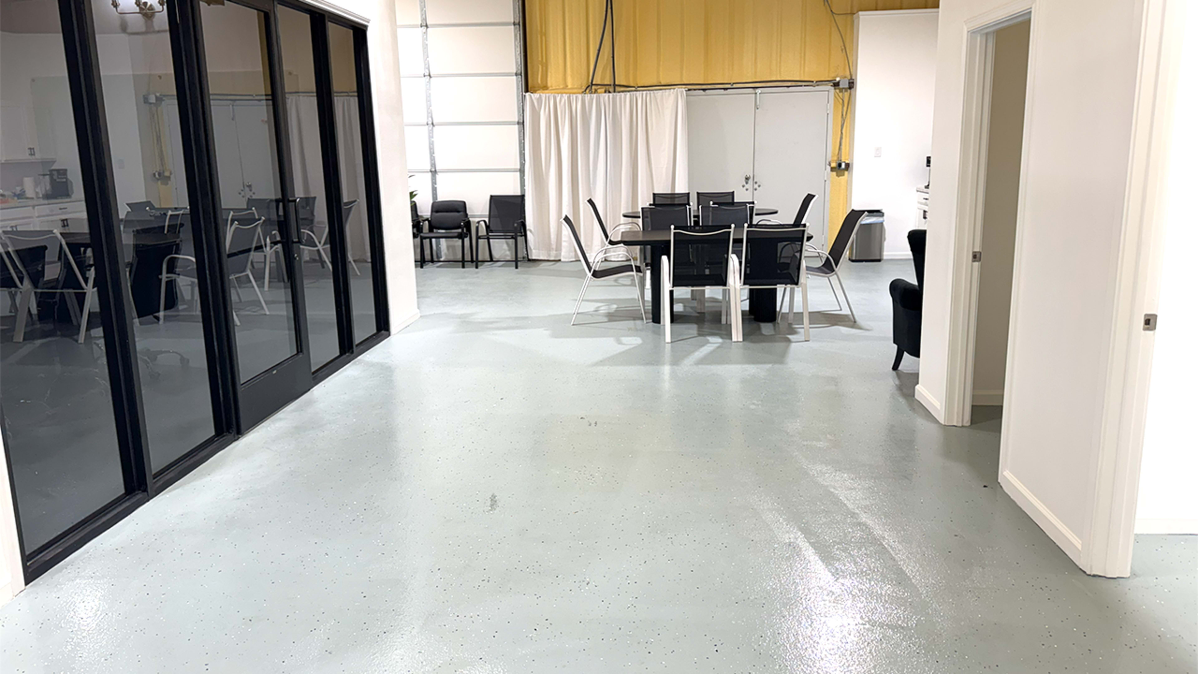 The image shows a modern indoor space with a polished concrete floor, featuring a dining table surrounded by chairs and large glass doors leading to an outdoor area.
