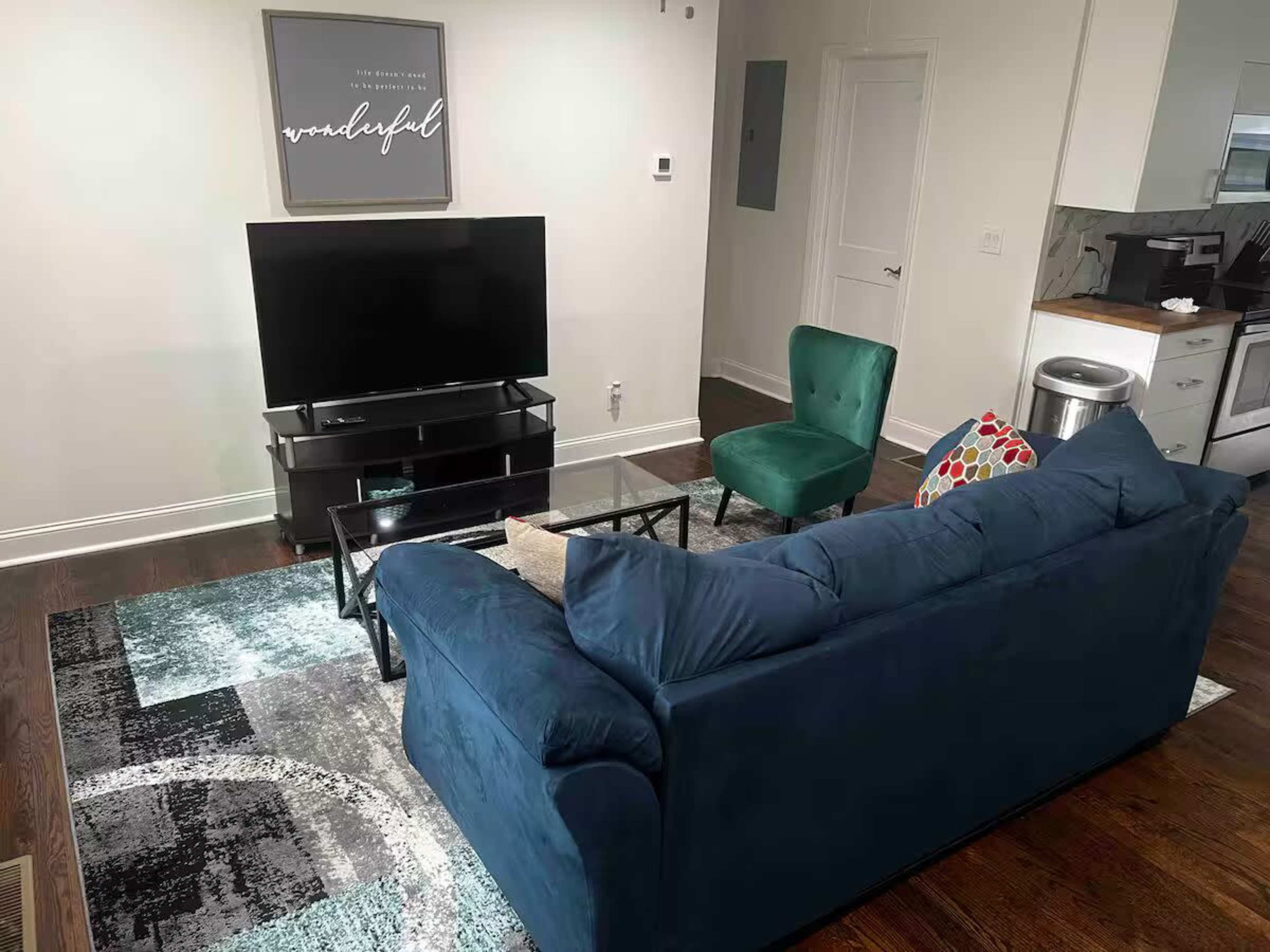 The image shows a living room with a dark blue couch facing a black television on a stand, alongside a green accent chair and a patterned rug on a hardwood floor.
