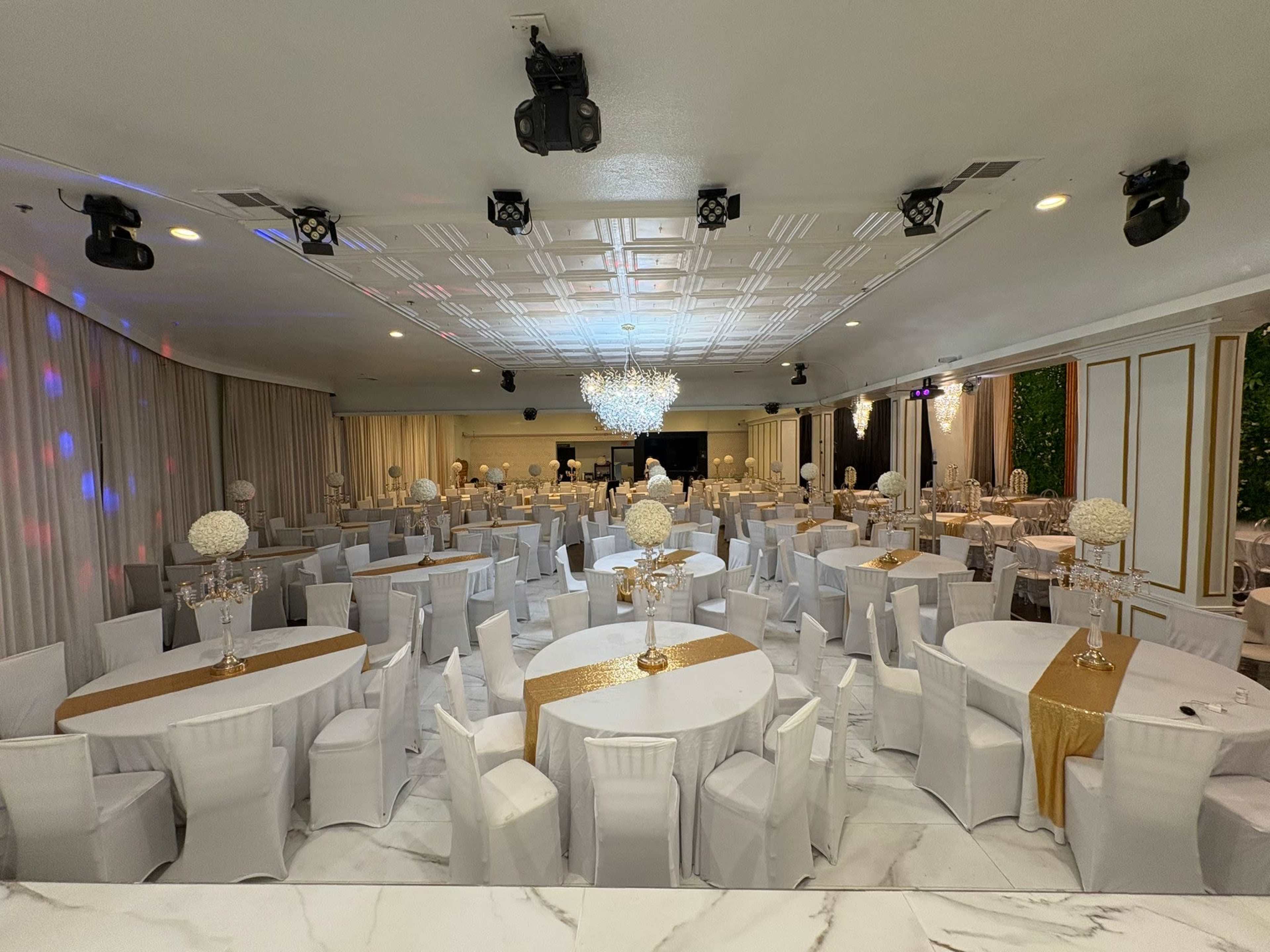 The image shows a large banquet hall decorated with white tables and chairs, elegant centerpieces, and subtle lighting fixtures.