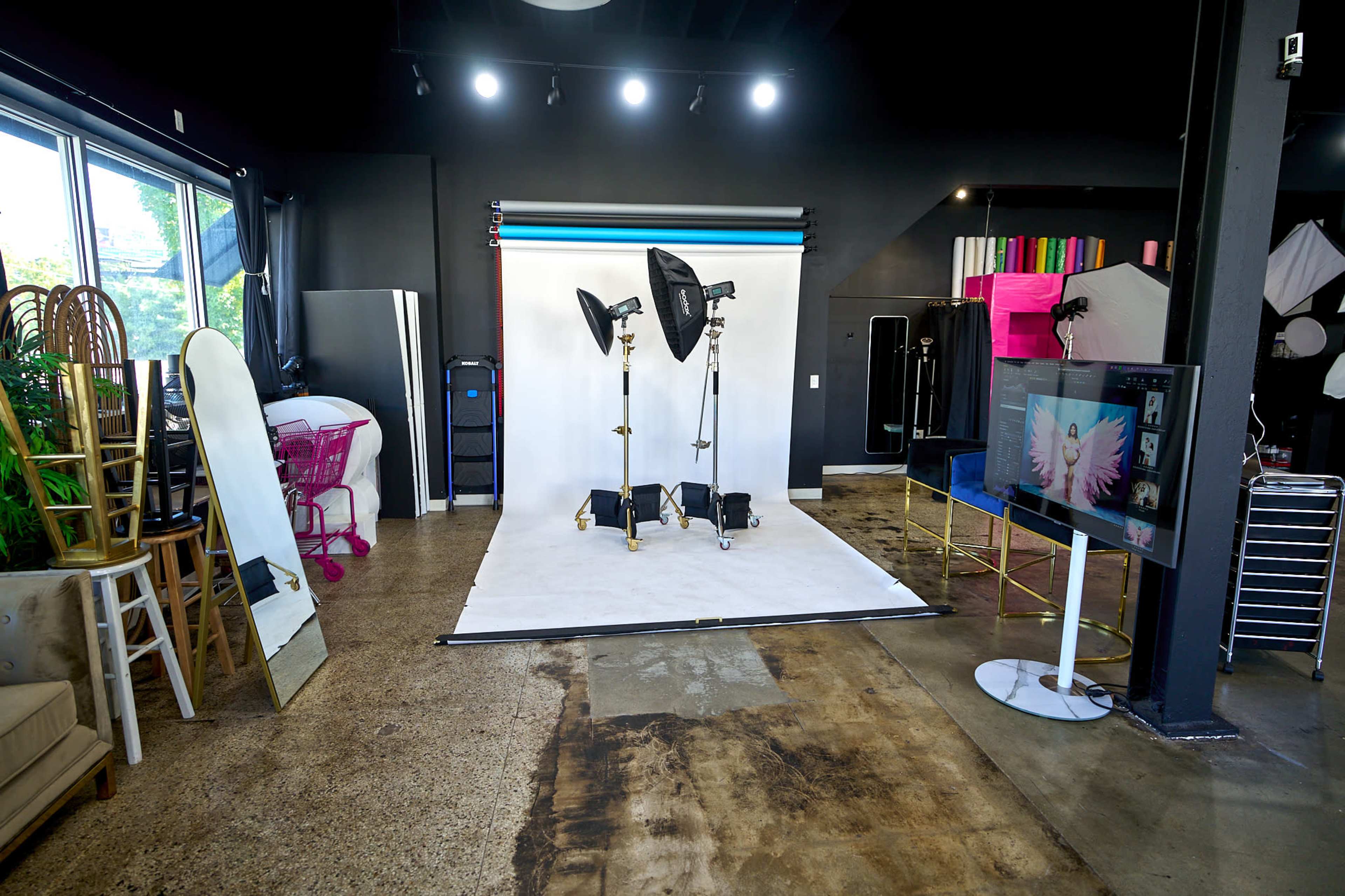A photography studio featuring a white backdrop, studio lights, a television, and various furniture pieces, including mirrors and chairs.