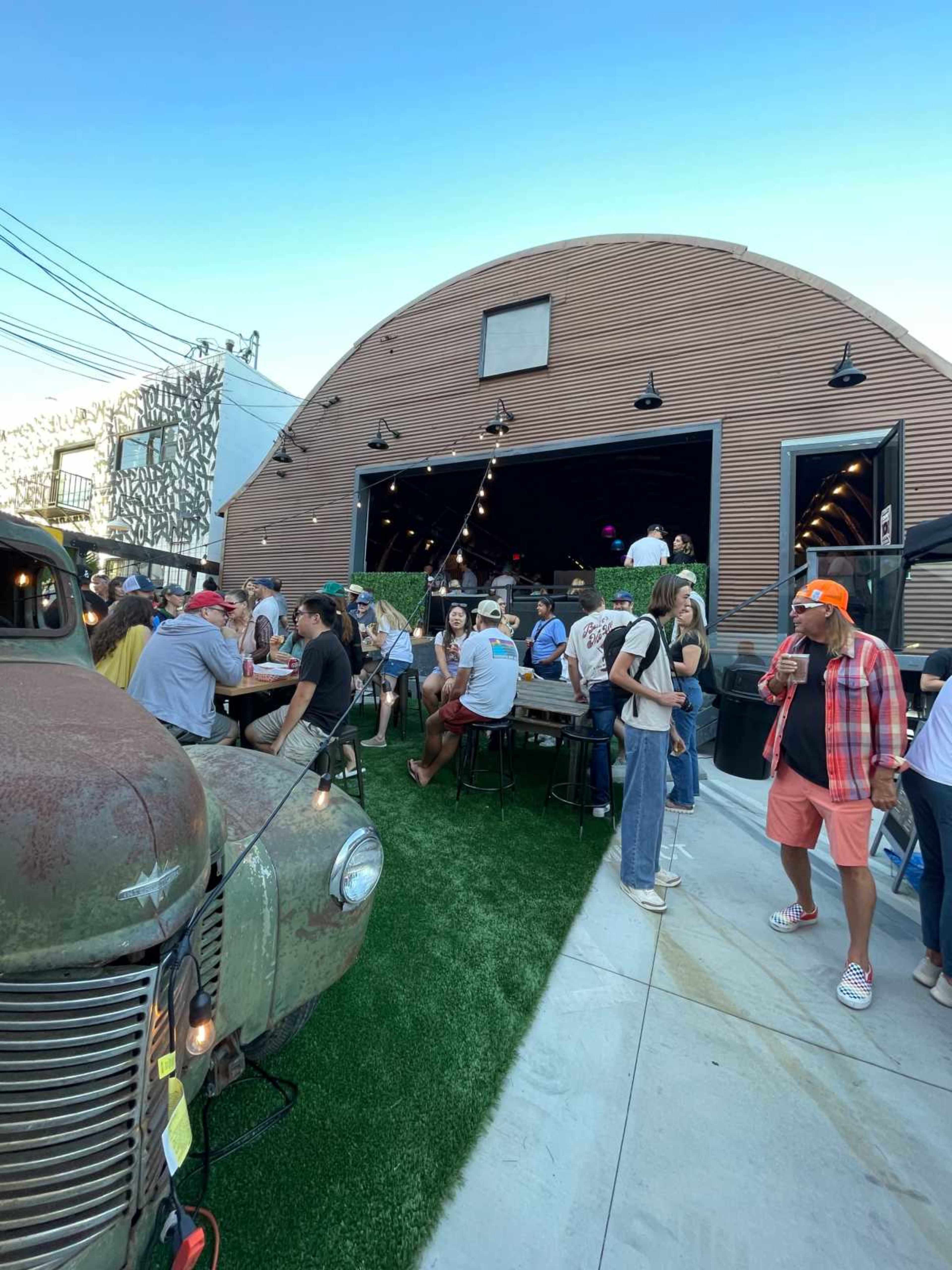 A crowd of people gathers around tables outside a large, modern building with a curved wooden facade, while a vintage truck is parked nearby.