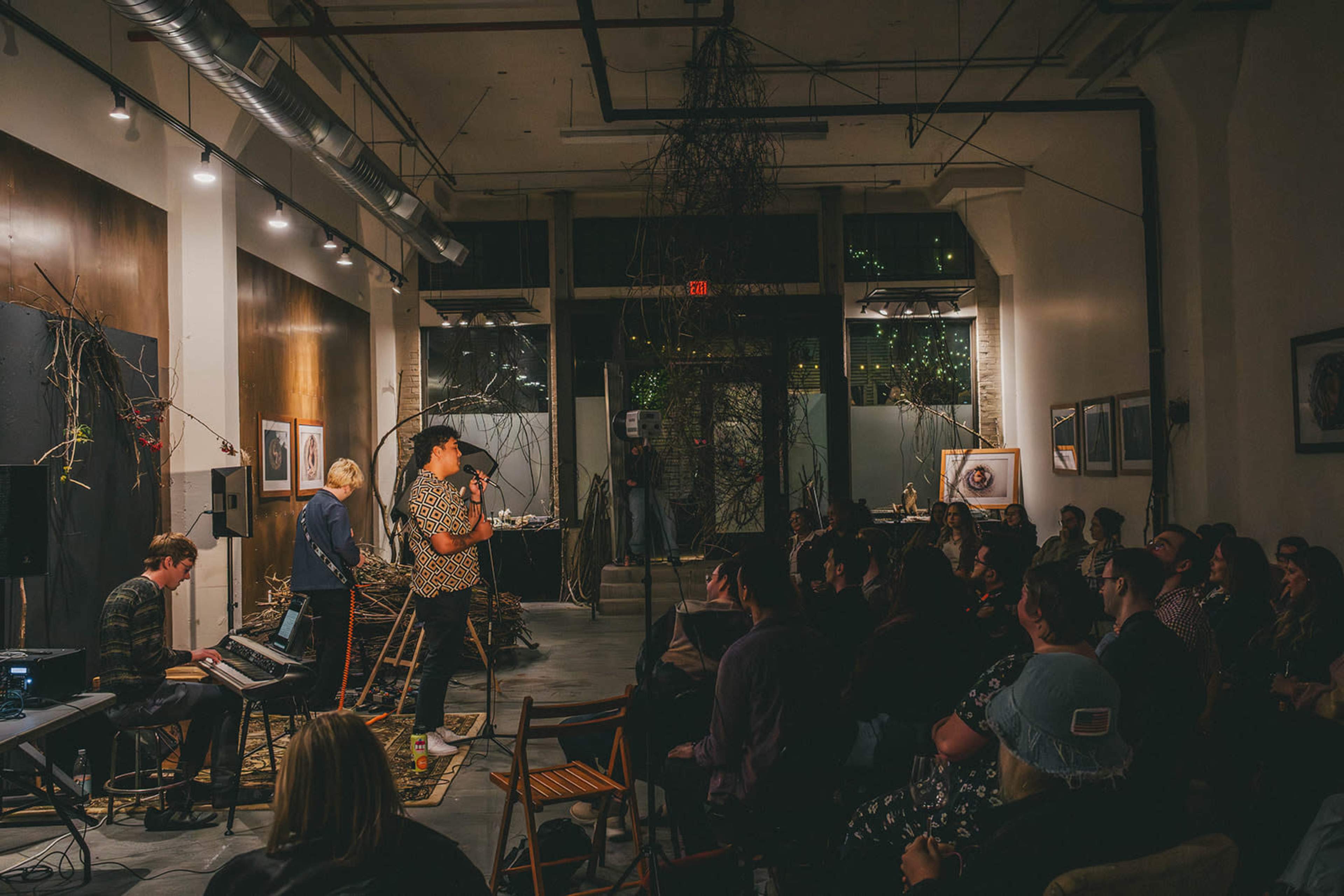 A crowd sits in an art gallery as a performer sings while accompanied by a keyboardist.