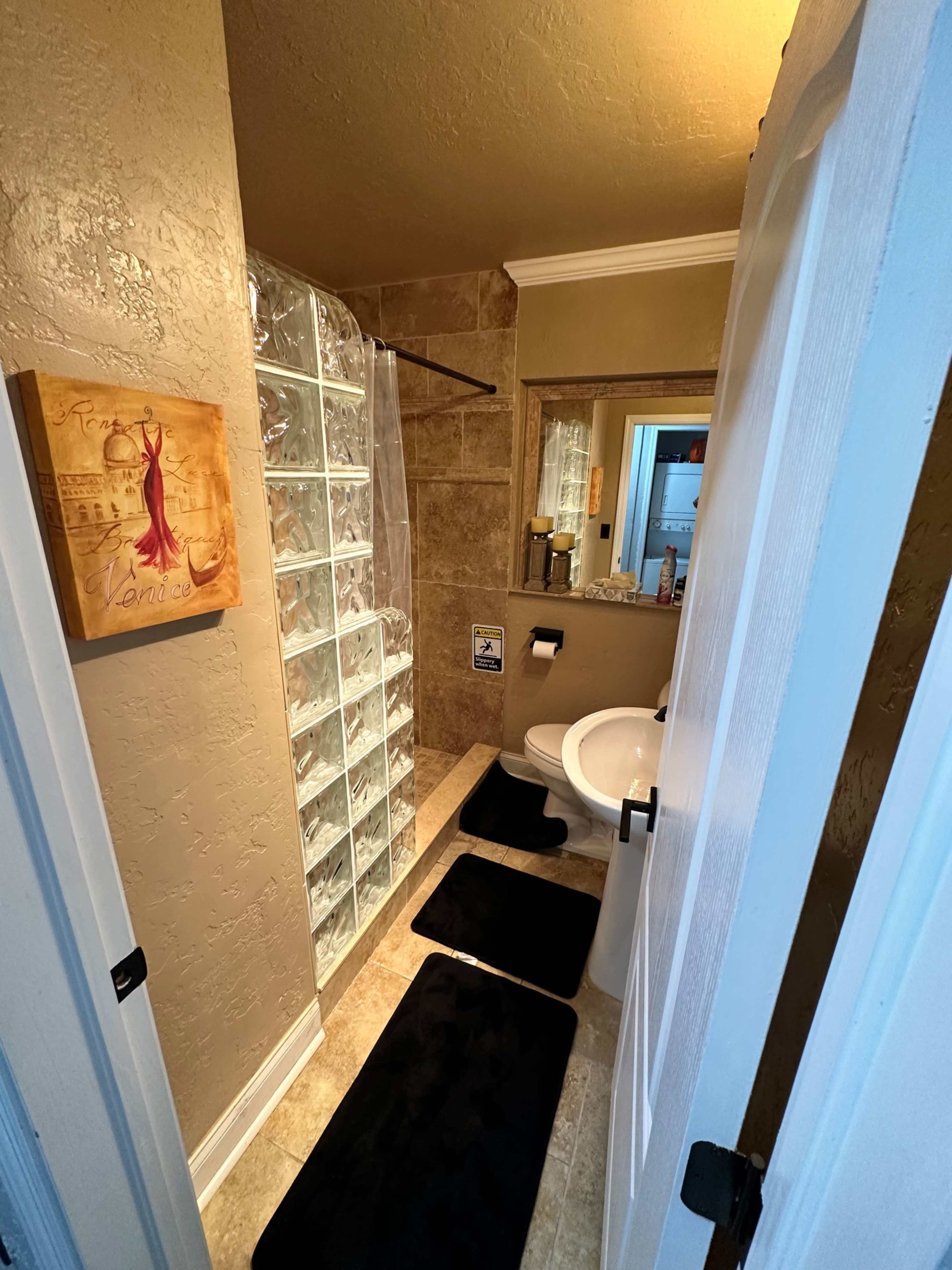 A narrow bathroom features a glass block shower, a white toilet, and two black bath mats on a tiled floor.