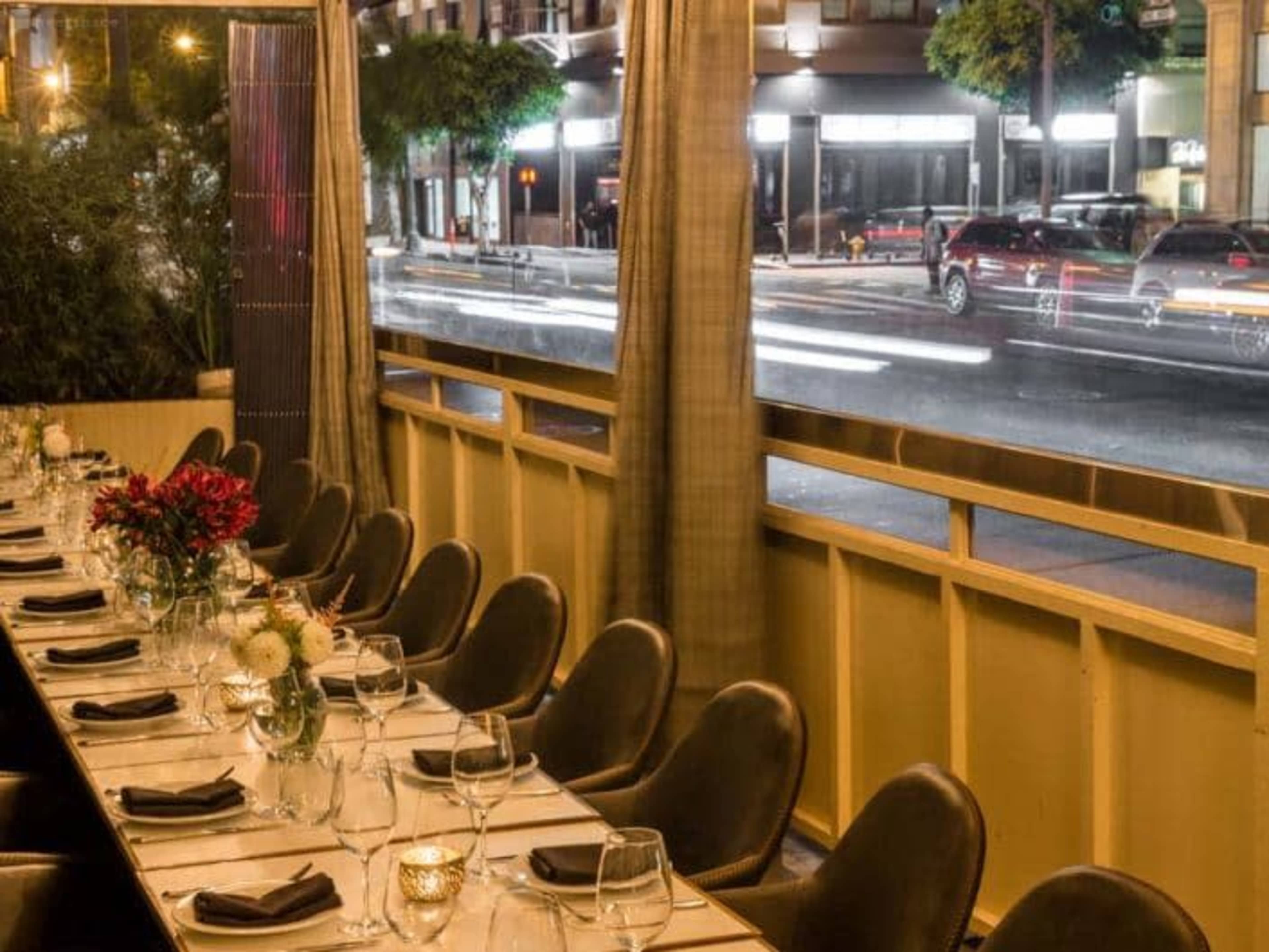 A long dining table is set with glasses and floral arrangements, positioned adjacent to a window overlooking a busy street at night.
