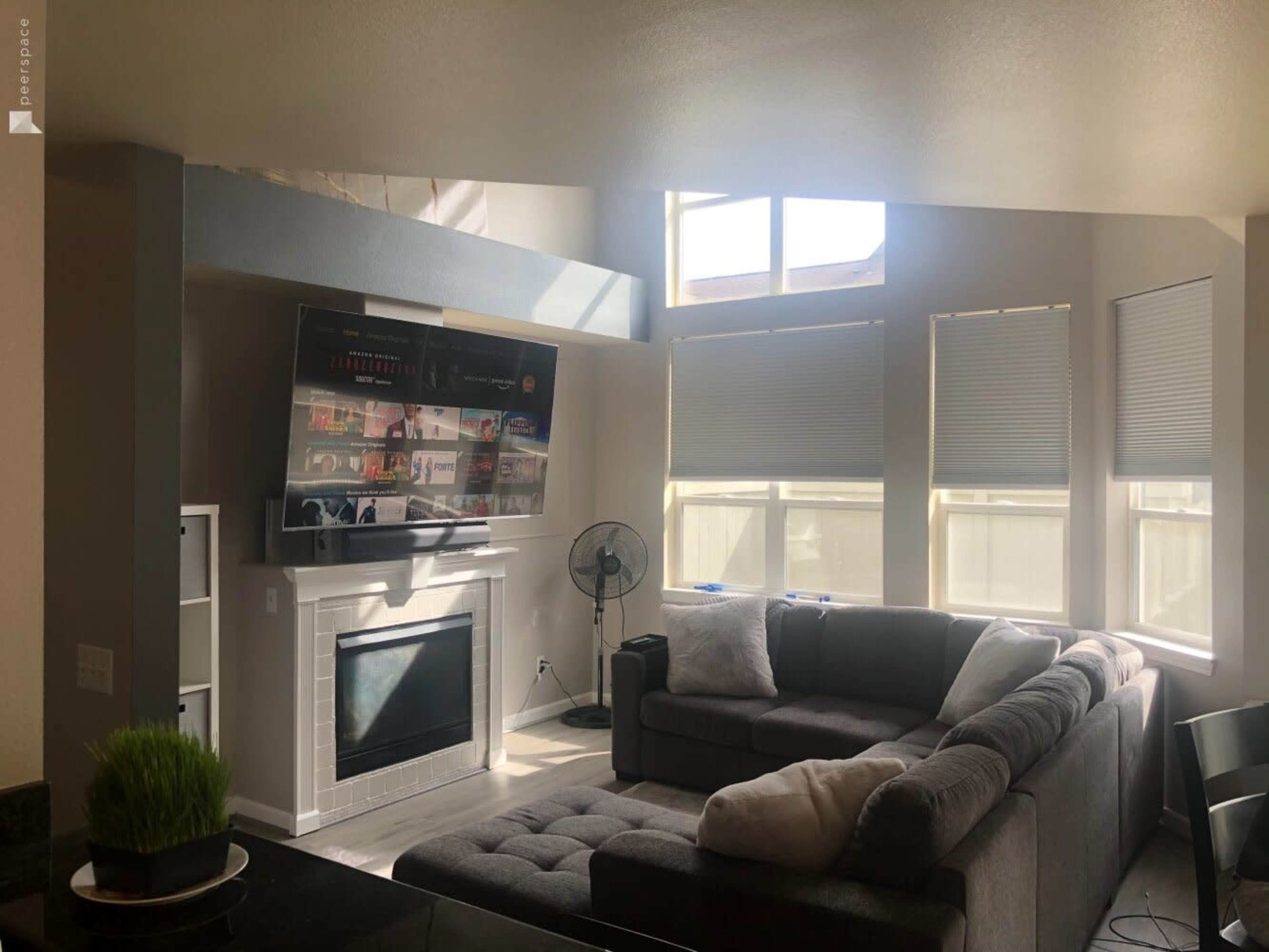 A modern living room with a large sectional sofa, a wall-mounted television, and bright windows allowing natural light to fill the space.