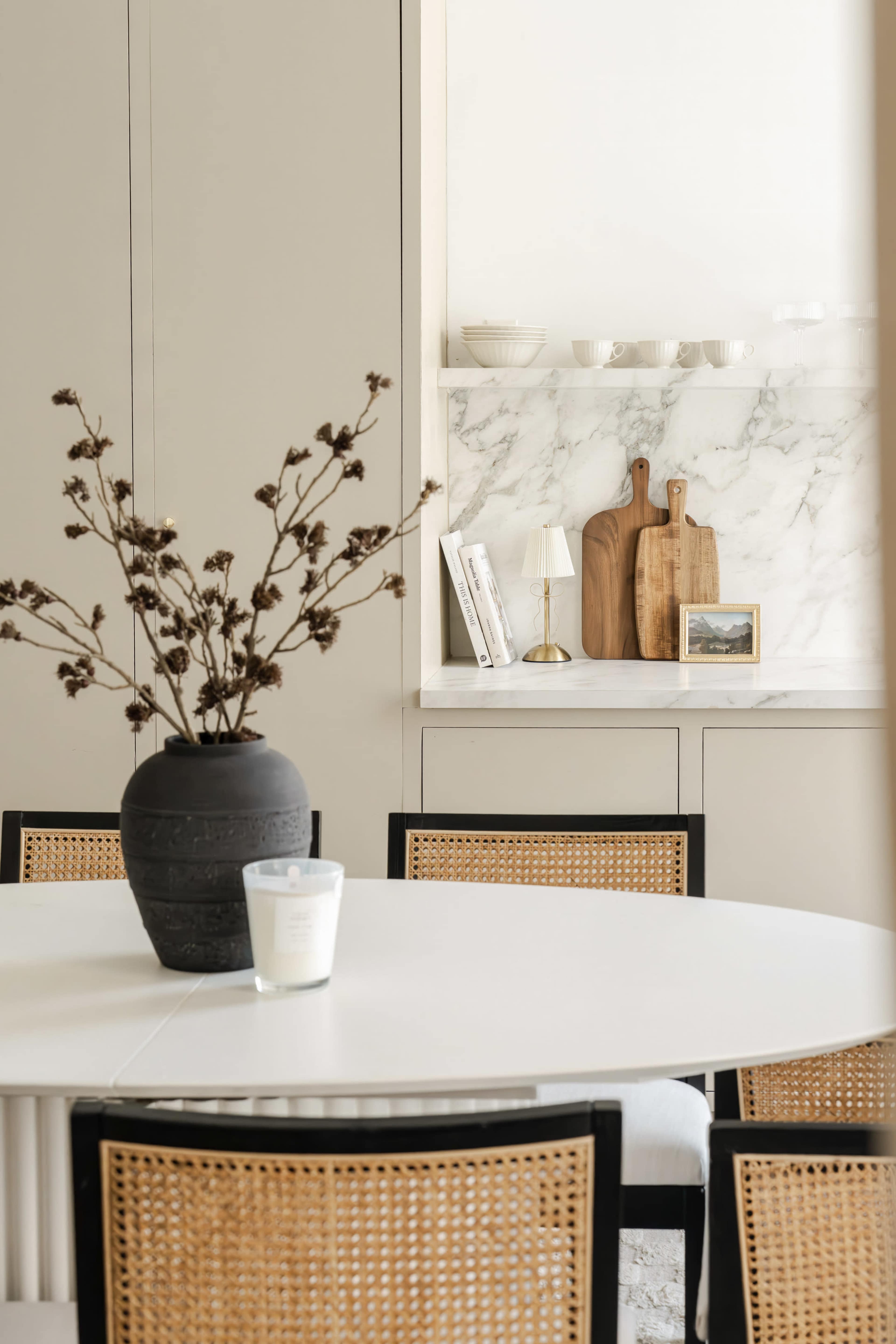 A modern dining area features a round white table with a black vase containing dried branches, surrounded by rattan chair seats, with a marble shelf displaying wooden cutting boards and a small lamp.