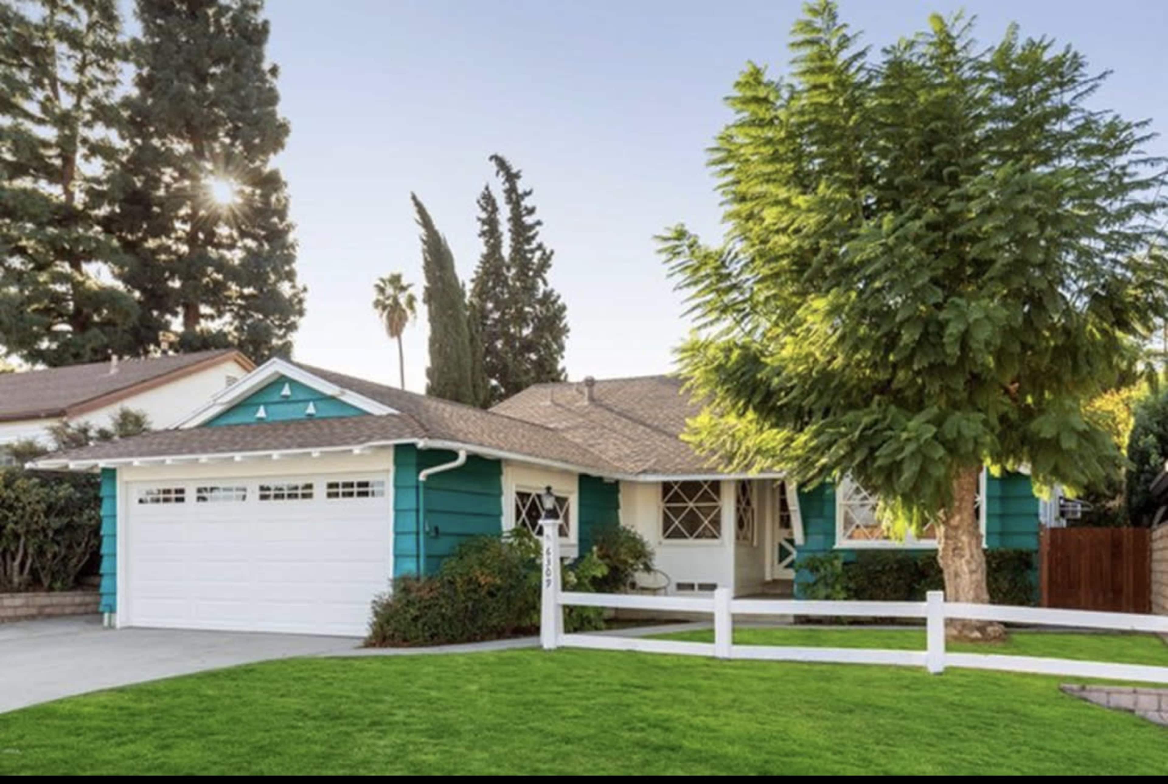 A teal-colored house with a garage and a white fence is surrounded by green grass and trees.