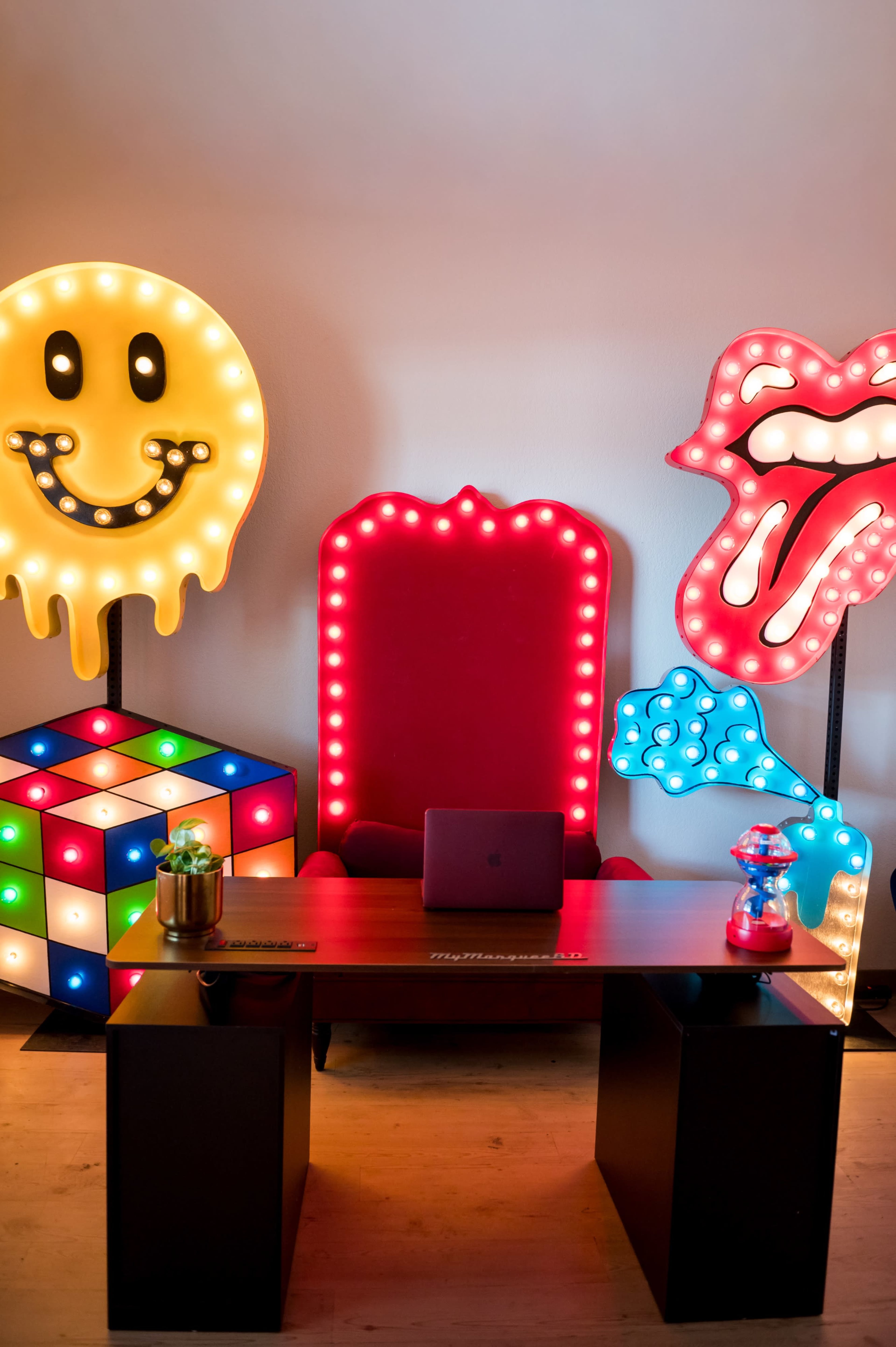 The image features a brightly lit room with a large red chair, colorful marquee signs of a smiling face and lips, a Rubik's Cube, and a desk holding a laptop and a decorative jar.