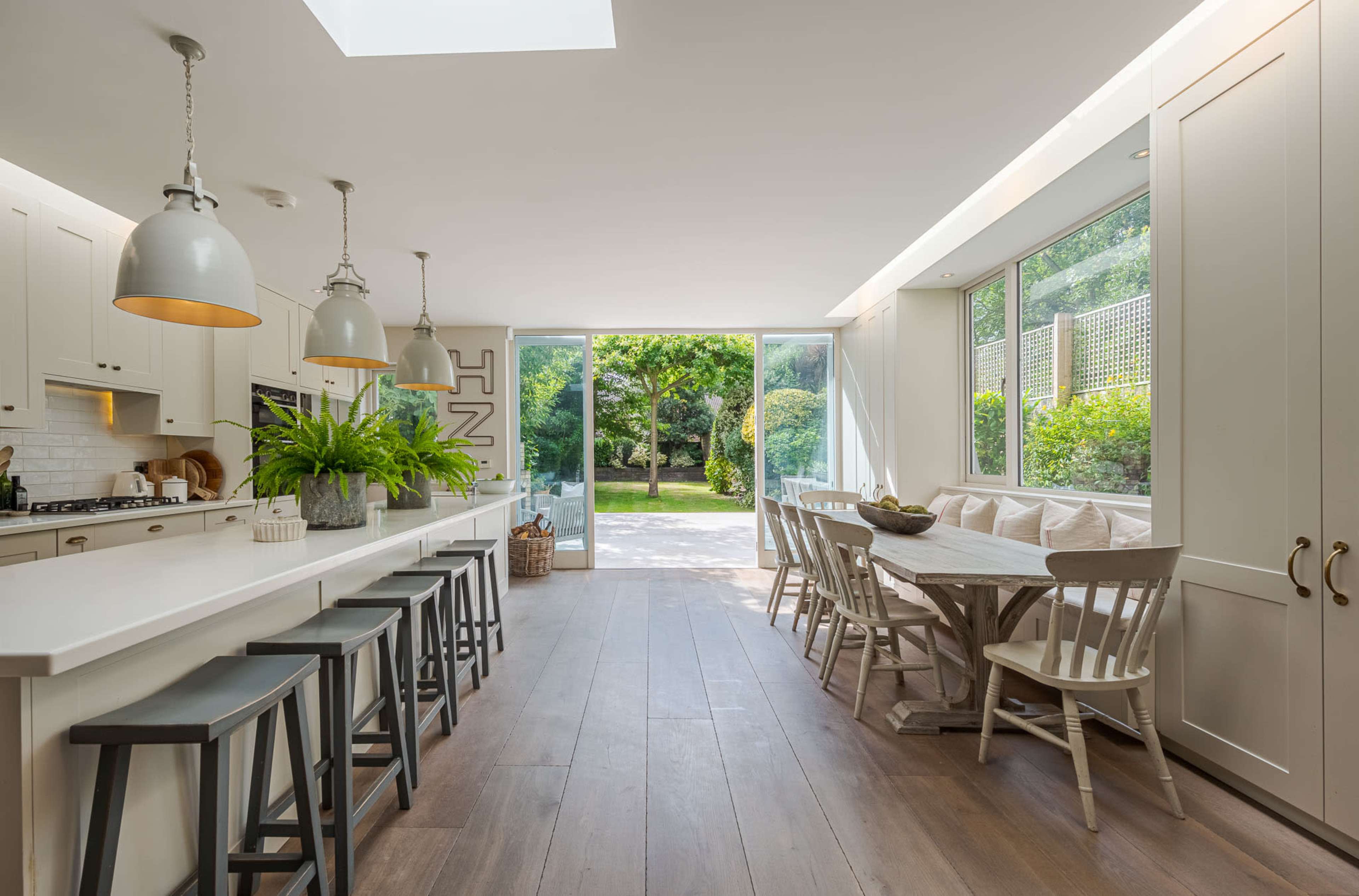 The image shows a modern kitchen and dining area with a large sliding glass door opening to a garden, featuring light-colored cabinetry, a central island with bar stools, and a wooden dining table surrounded by chairs.
