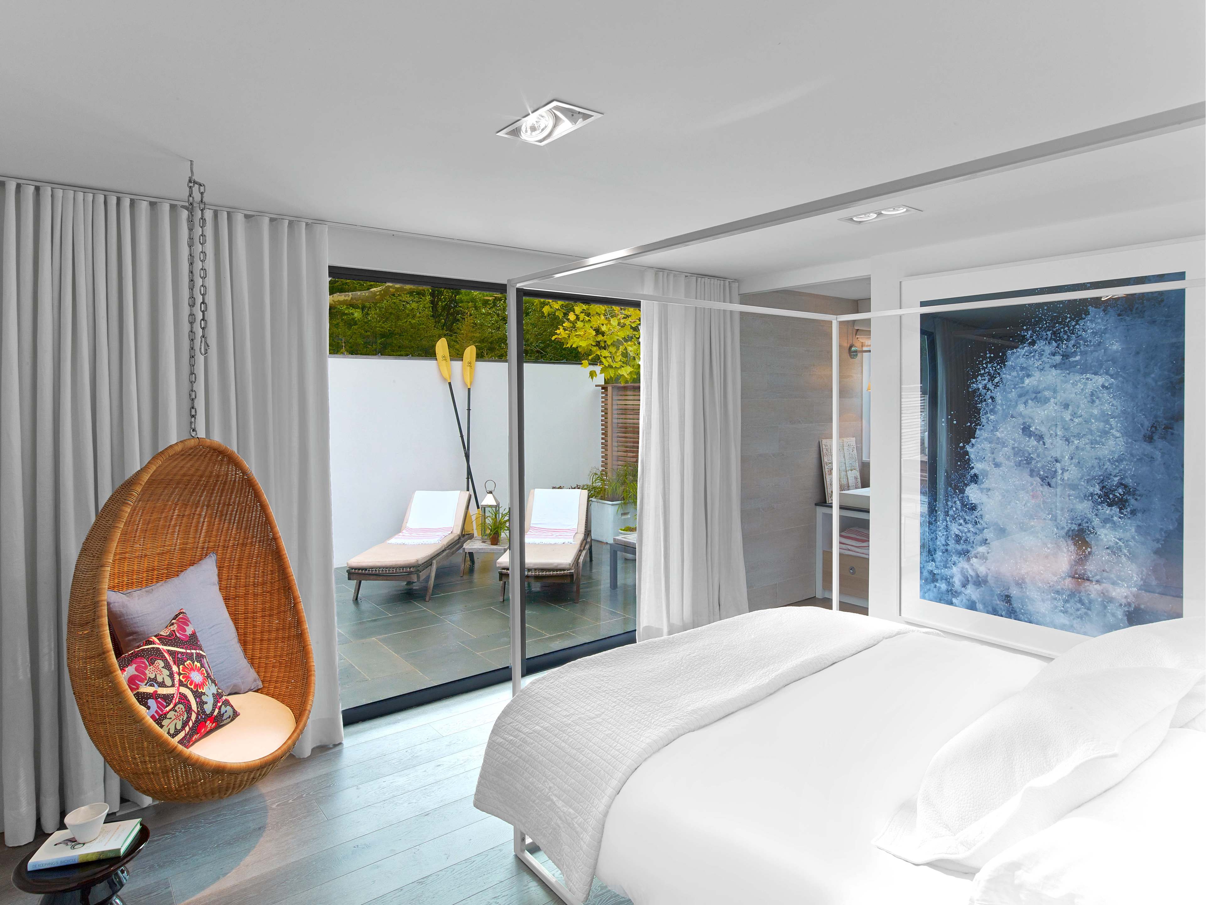A modern bedroom features a hanging wicker chair and glass doors leading to a patio with two sun loungers.