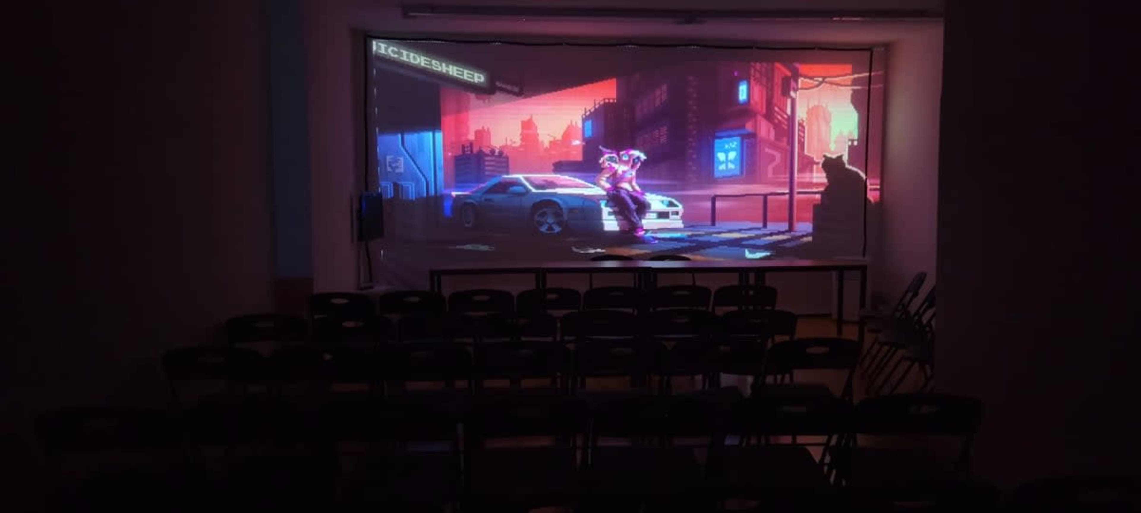A colorful digital art scene depicting a futuristic city is projected onto a screen in a dimly lit room with black chairs arranged in rows.