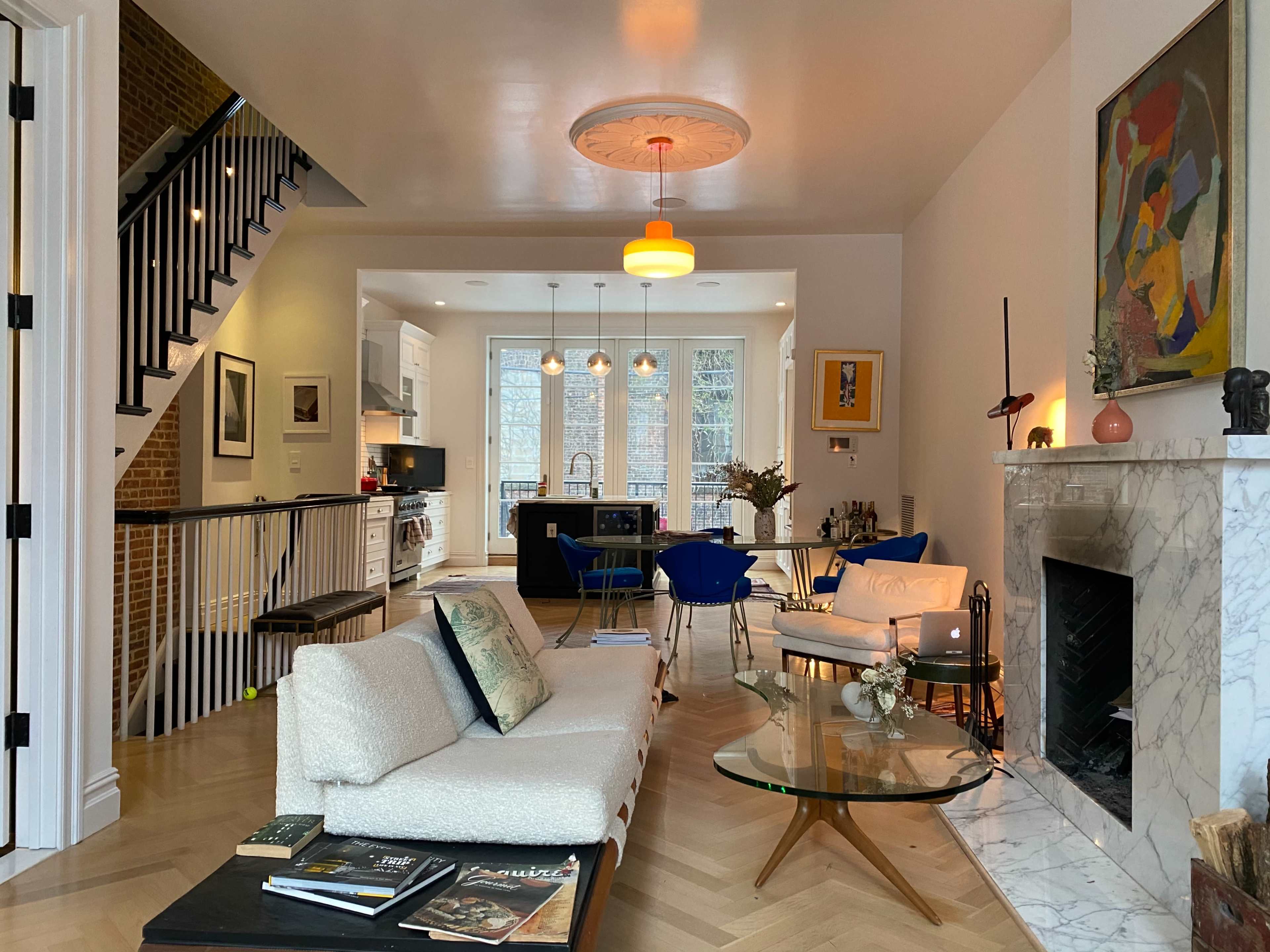 The image shows a contemporary living space featuring a white sofa, a glass coffee table, and a staircase leading to another level, with an open kitchen visible in the background.