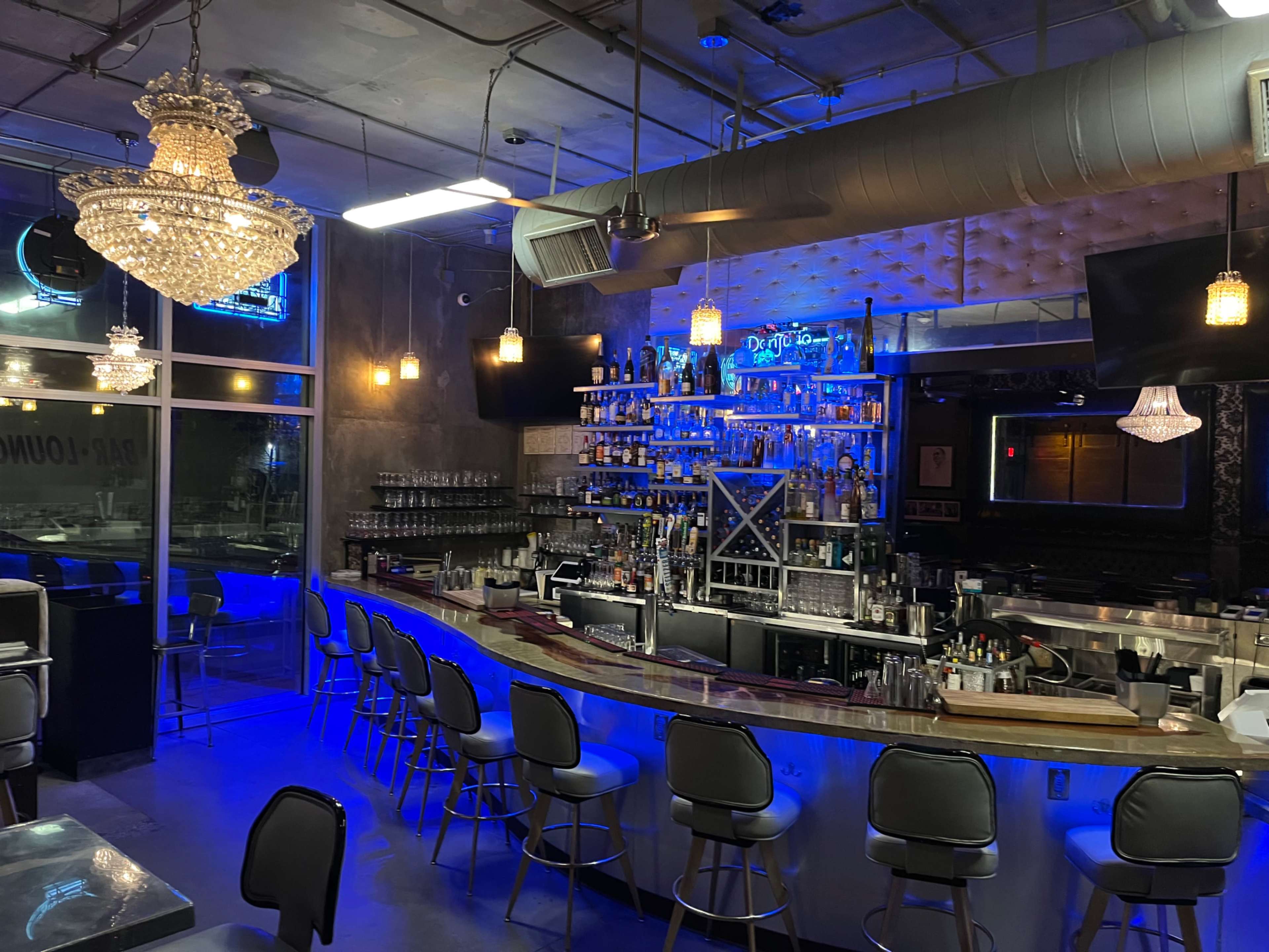 A modern bar interior featuring a curved bar with a variety of liquor bottles, stylish lighting fixtures, and high stools arranged around the counter.