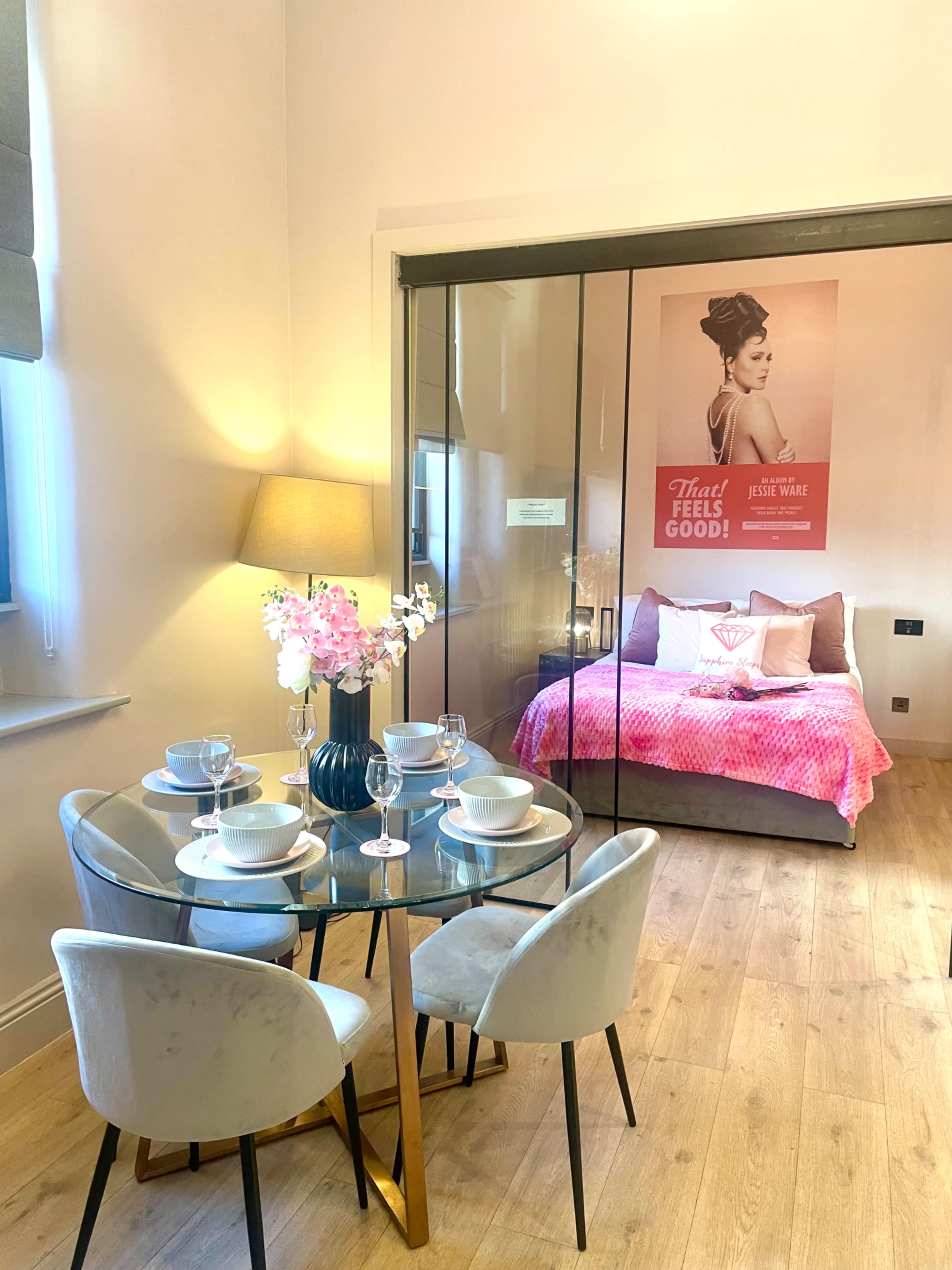 A modern dining area features a circular glass table with white chairs, overlooking a bedroom enclosed in glass with a large poster on the wall.
