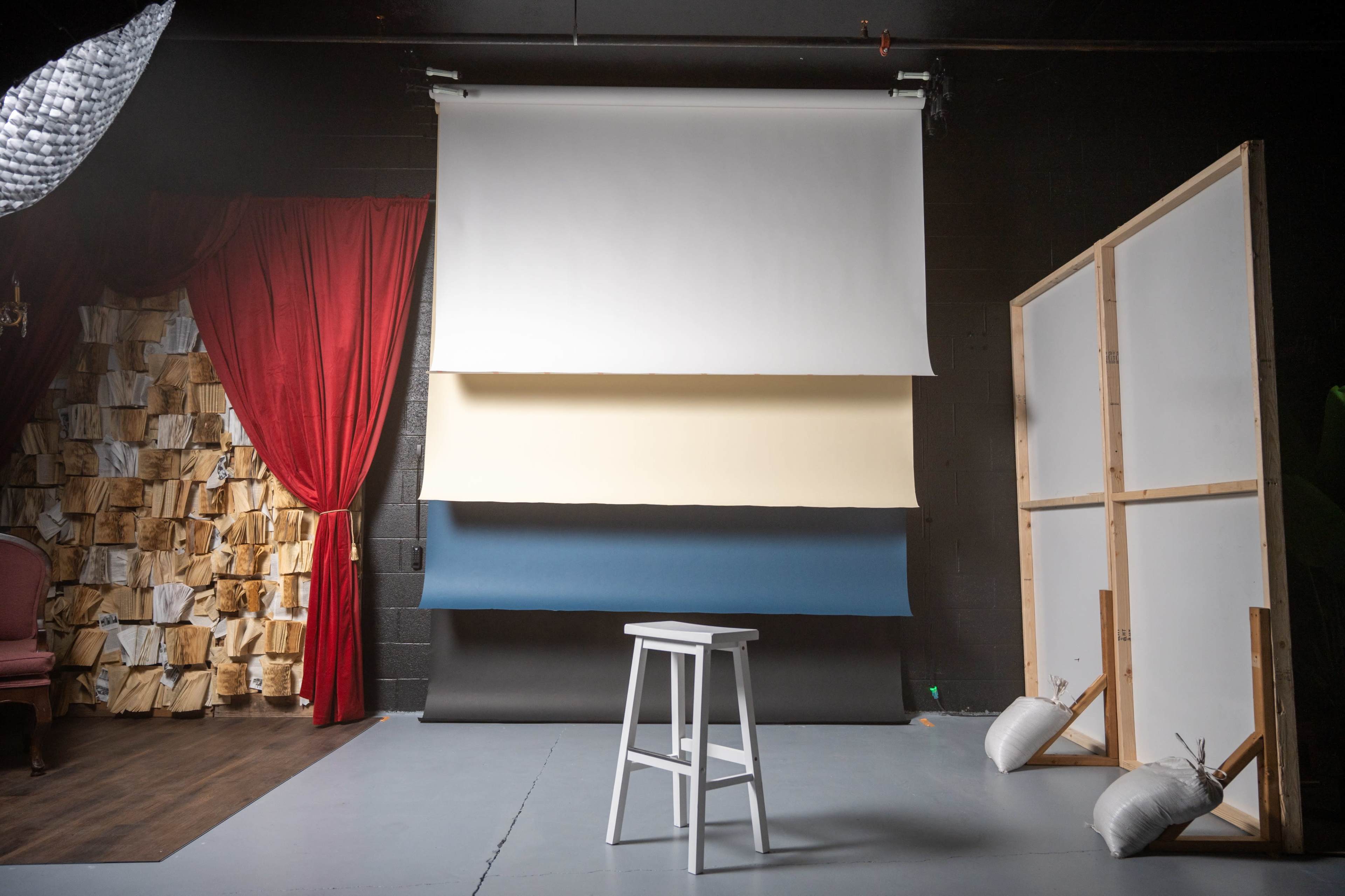 The image shows a photography studio with a white stool in front of three backdrop roll selections and a red curtain on one side.