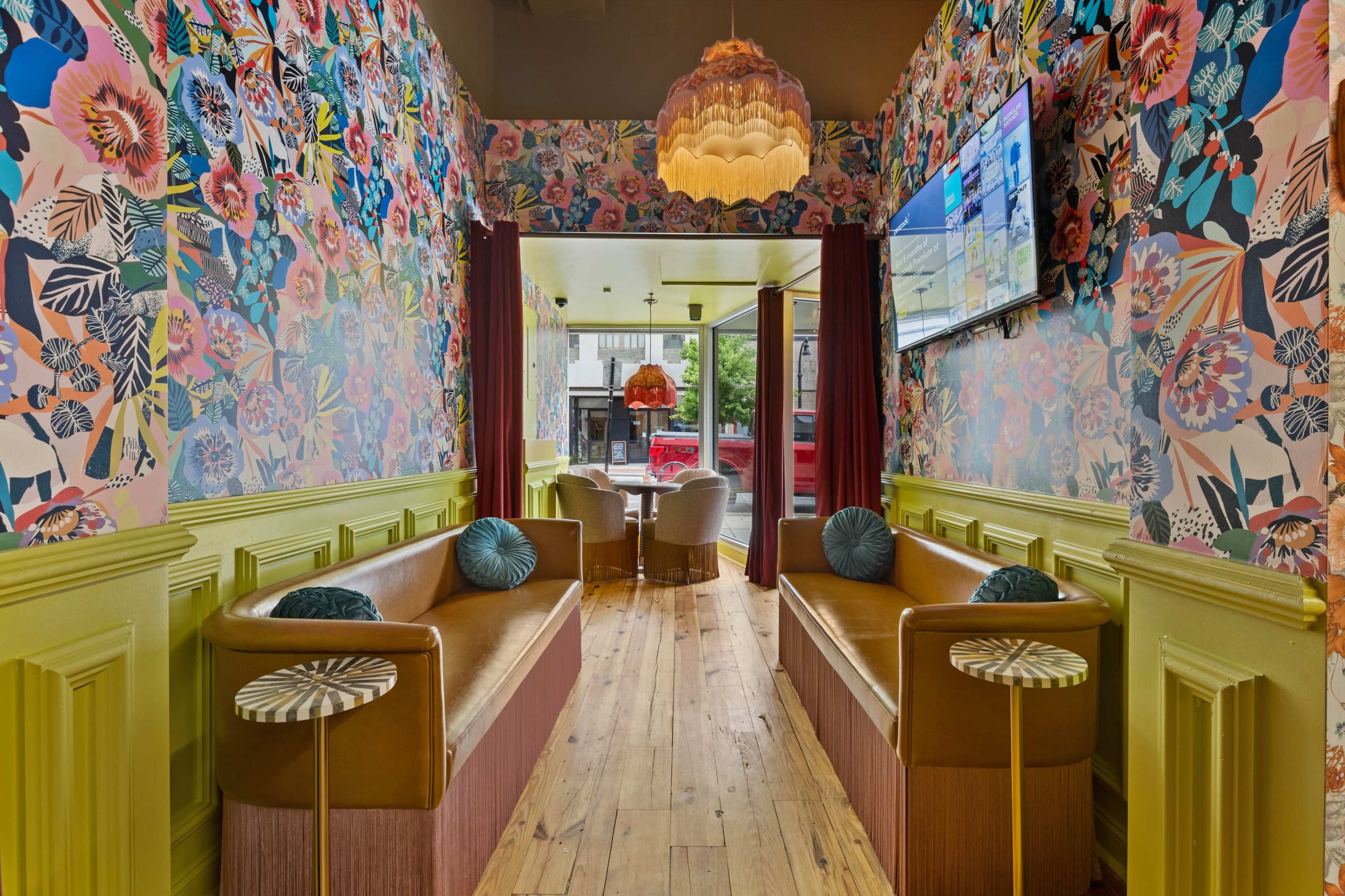 The image shows a hallway with vibrant floral wallpaper, two brown sofas, and a round table with chairs at the far end, illuminated by natural light.