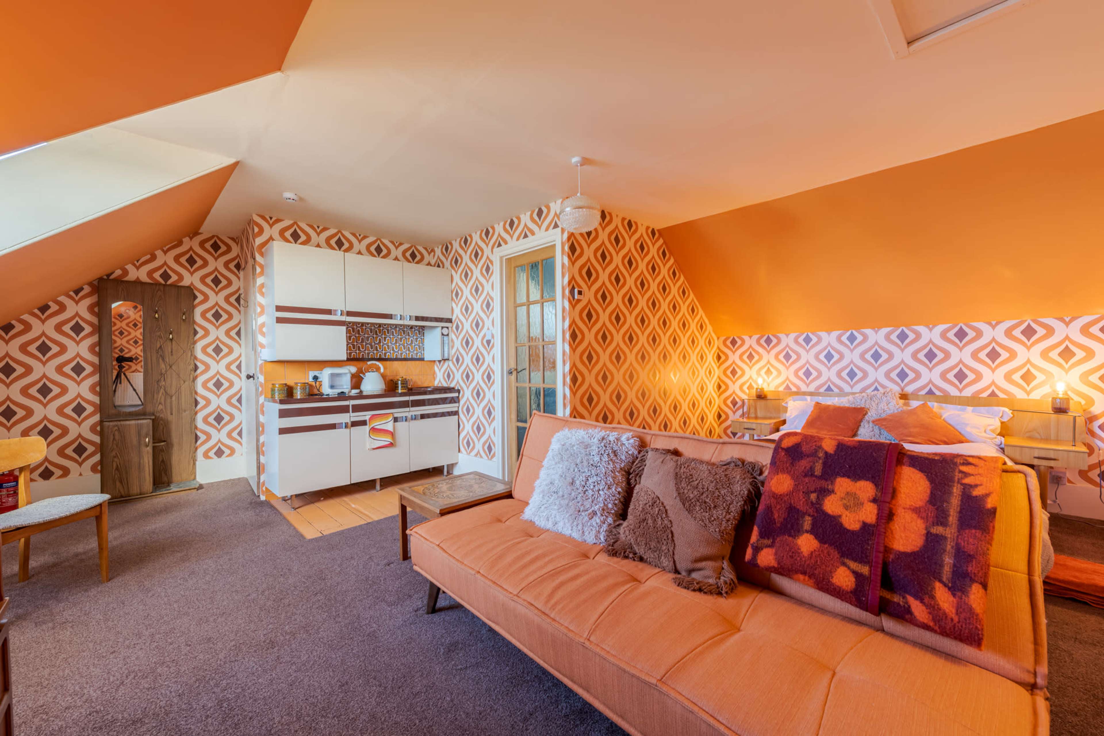 A cozy attic apartment features an orange color scheme with patterned wallpaper, a kitchenette, and a sofa adorned with decorative pillows.
