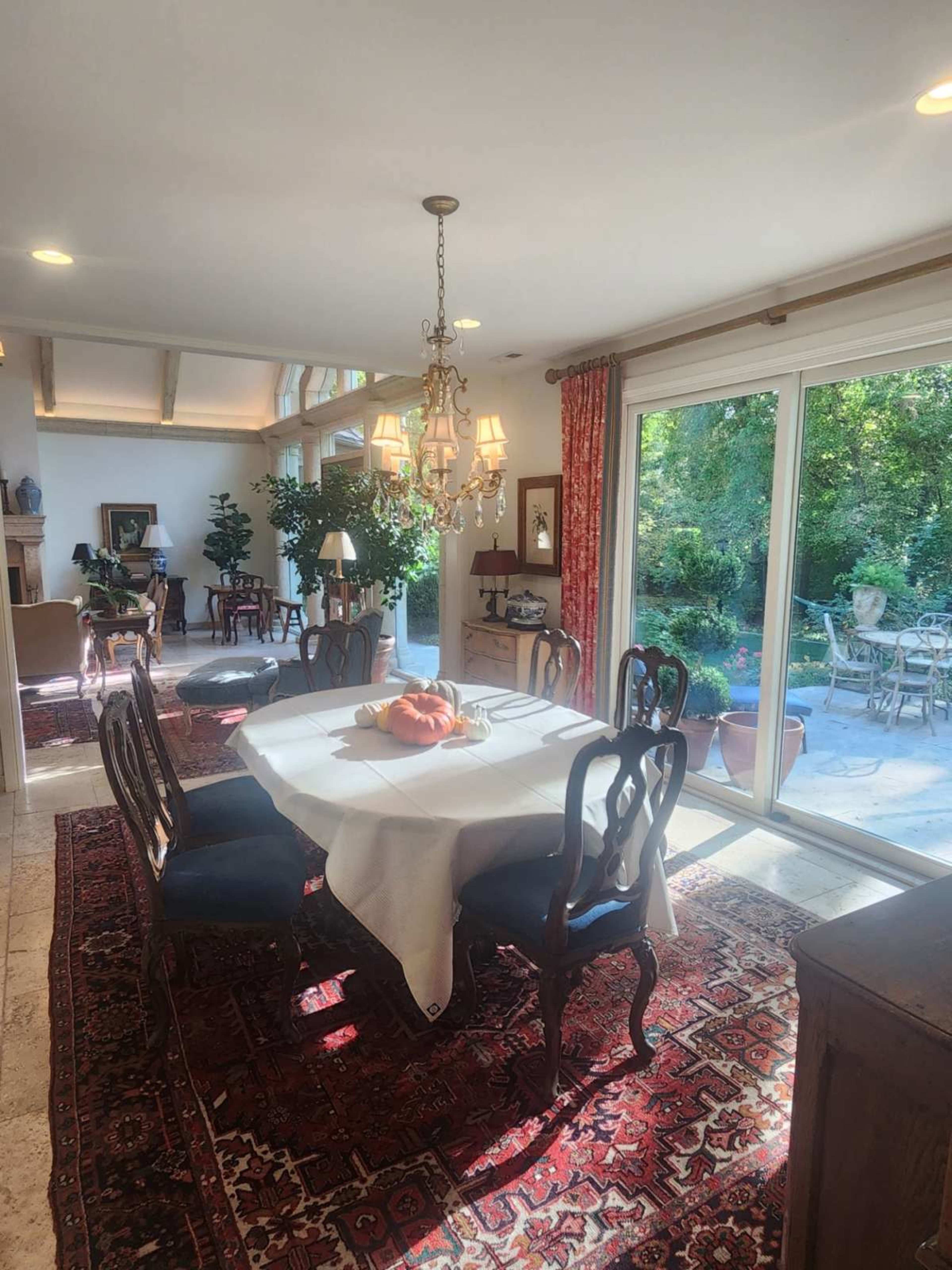 The image shows a dining area with a round table set for six, surrounded by elegant chairs, and large windows revealing a garden view.