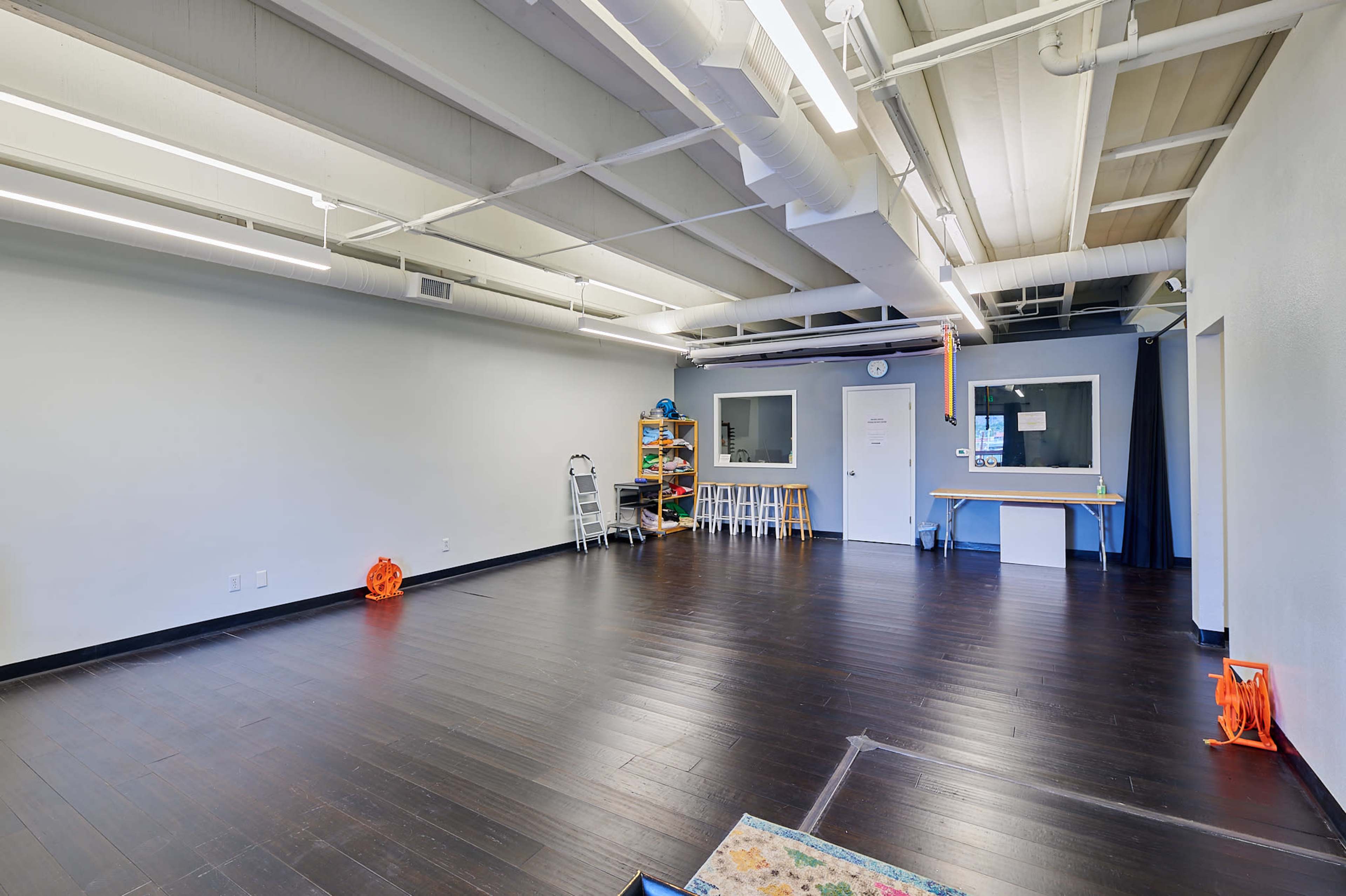 The image shows a spacious, empty room with brown wooden flooring, white walls, and a few pieces of furniture against the walls.