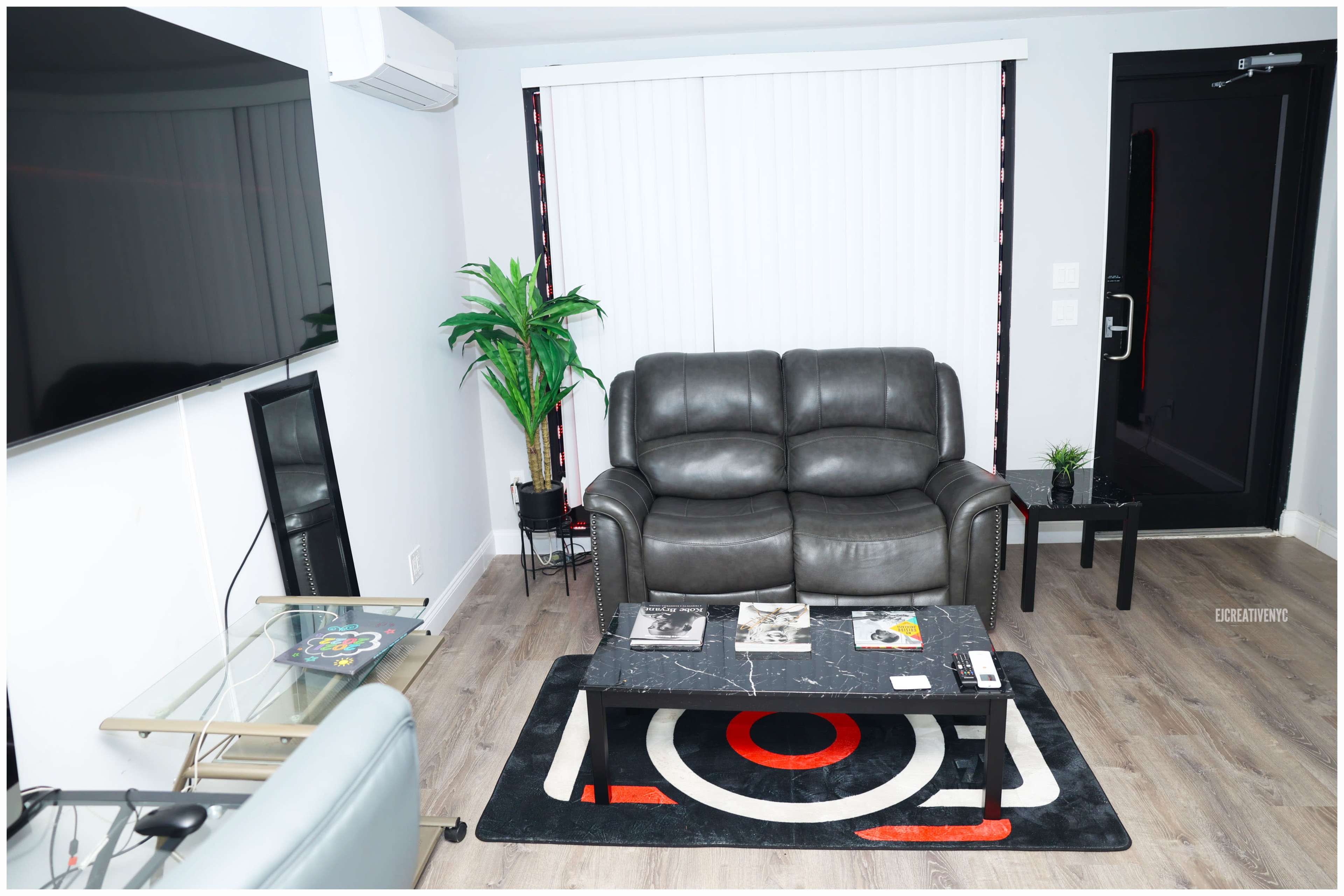 A living room with a gray leather loveseat, a black television mounted on the wall, a small table, and a decorative plant near a window with vertical blinds.