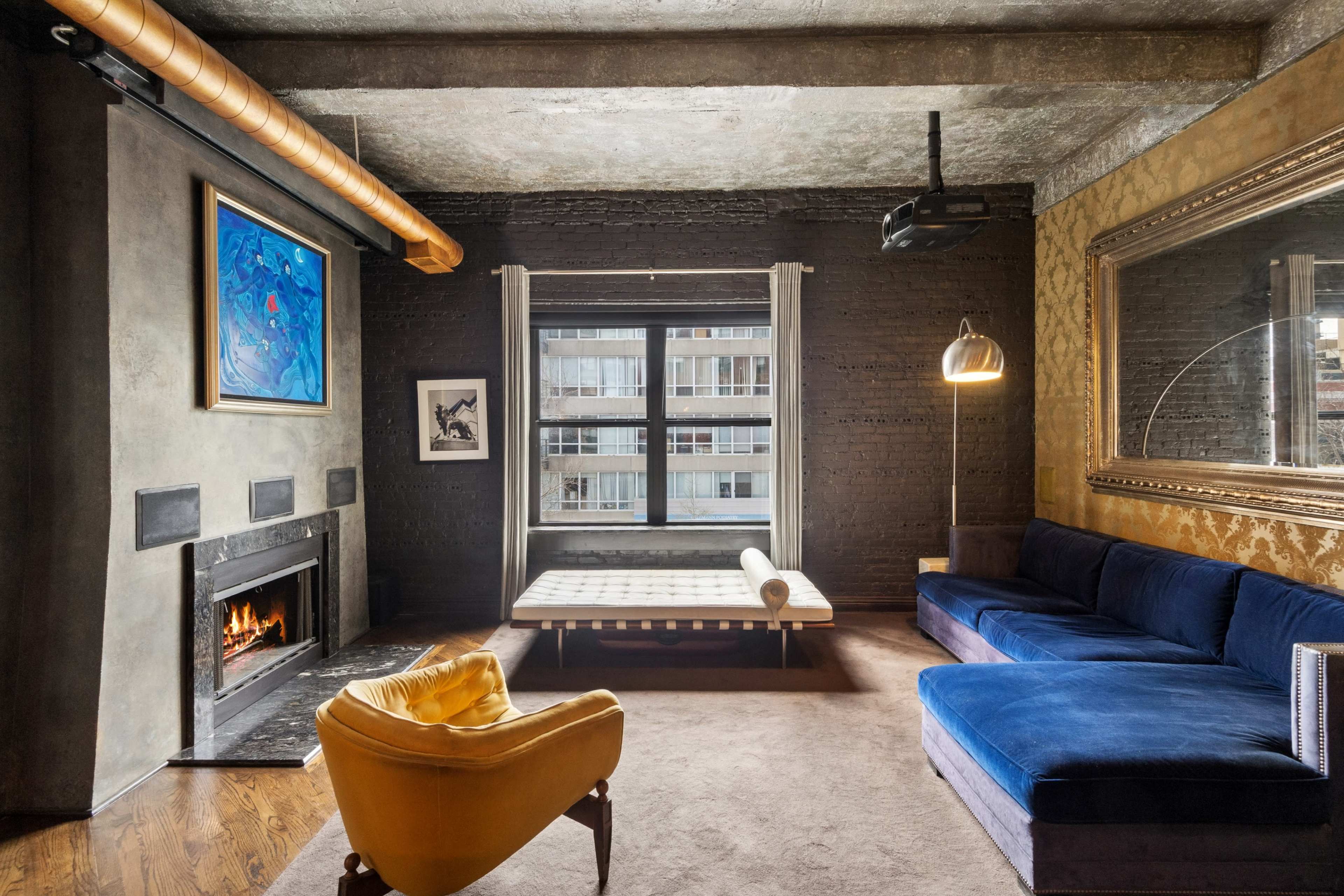 The image shows a modern living room with a blue sectional sofa, a yellow armchair, and a fireplace, framed by large windows and decorative walls.