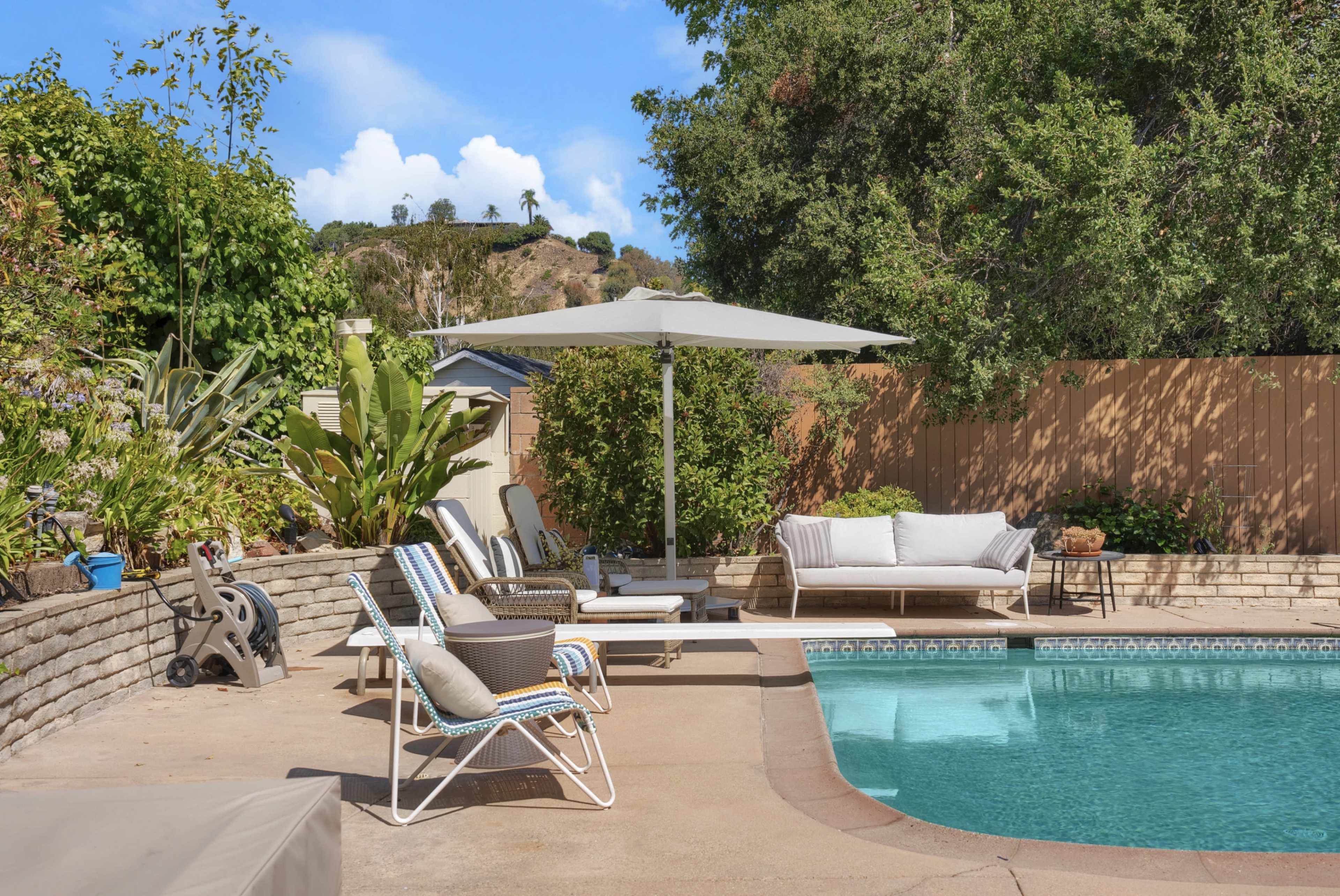 The image shows a residential pool area featuring lounge chairs, an umbrella, and surrounding greenery.