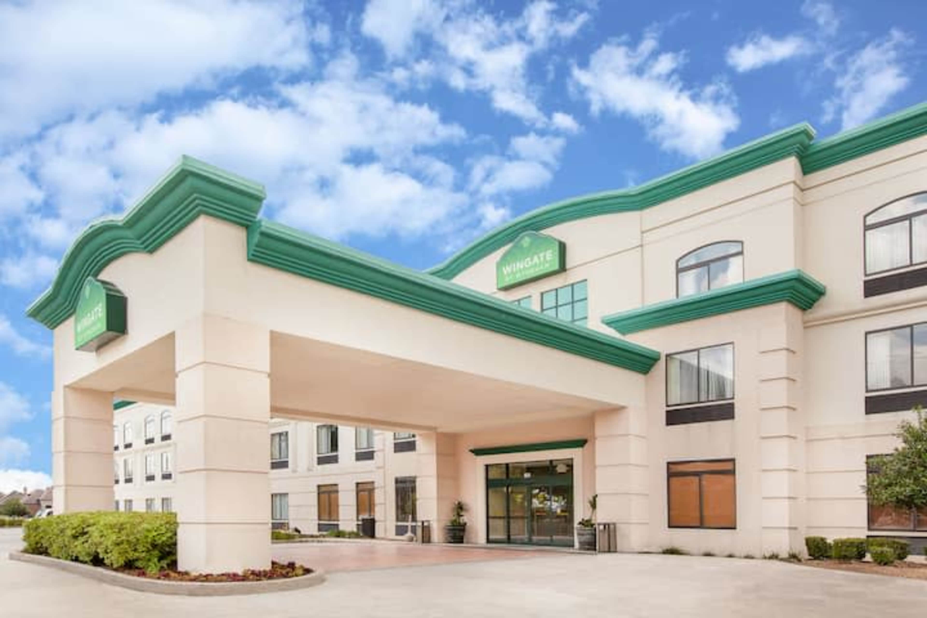The entrance of a Wingate hotel building with a pale exterior and a green accent roof under a blue sky.