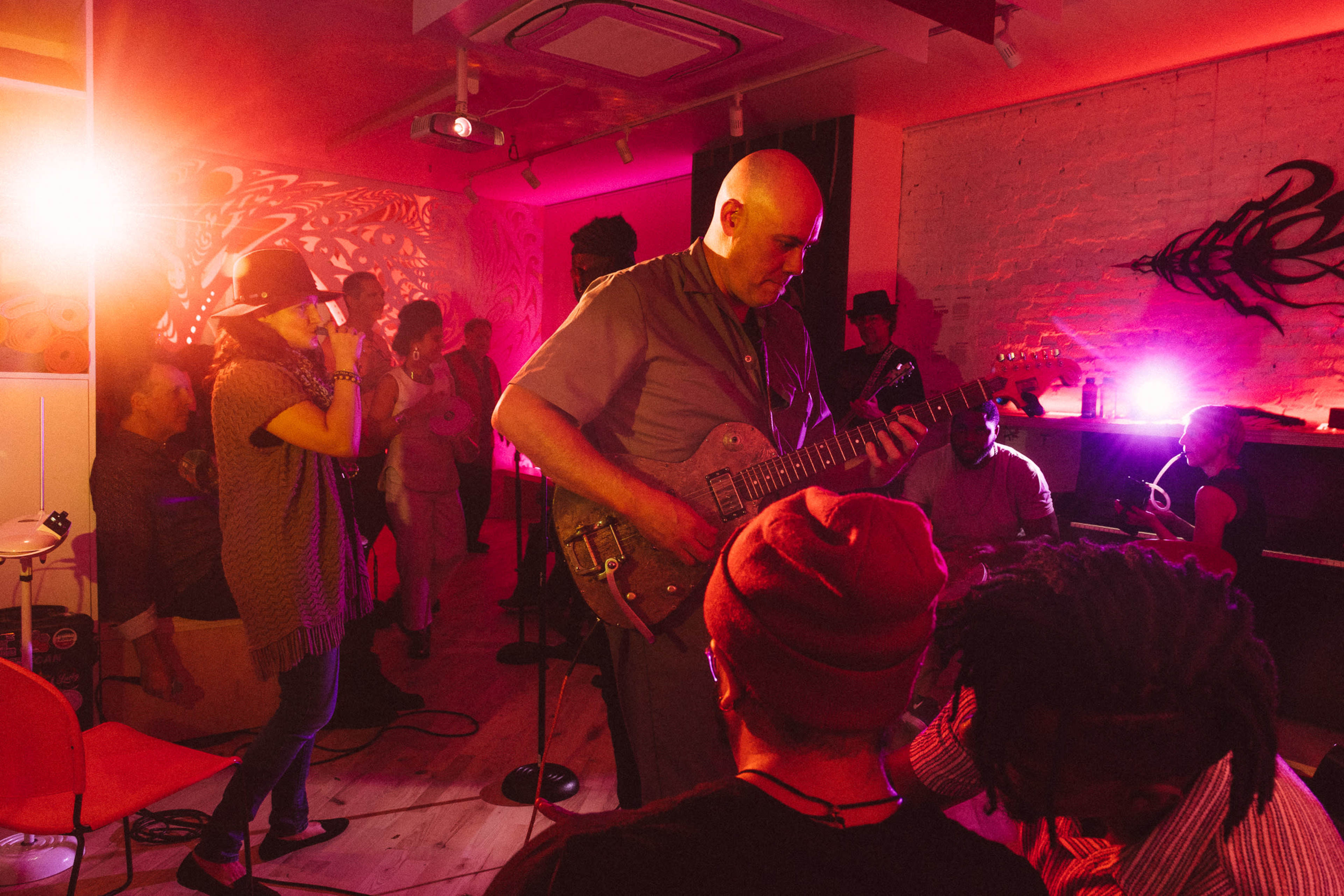 A musician plays guitar while people gather and enjoy a live music performance in a vibrant, illuminated room.