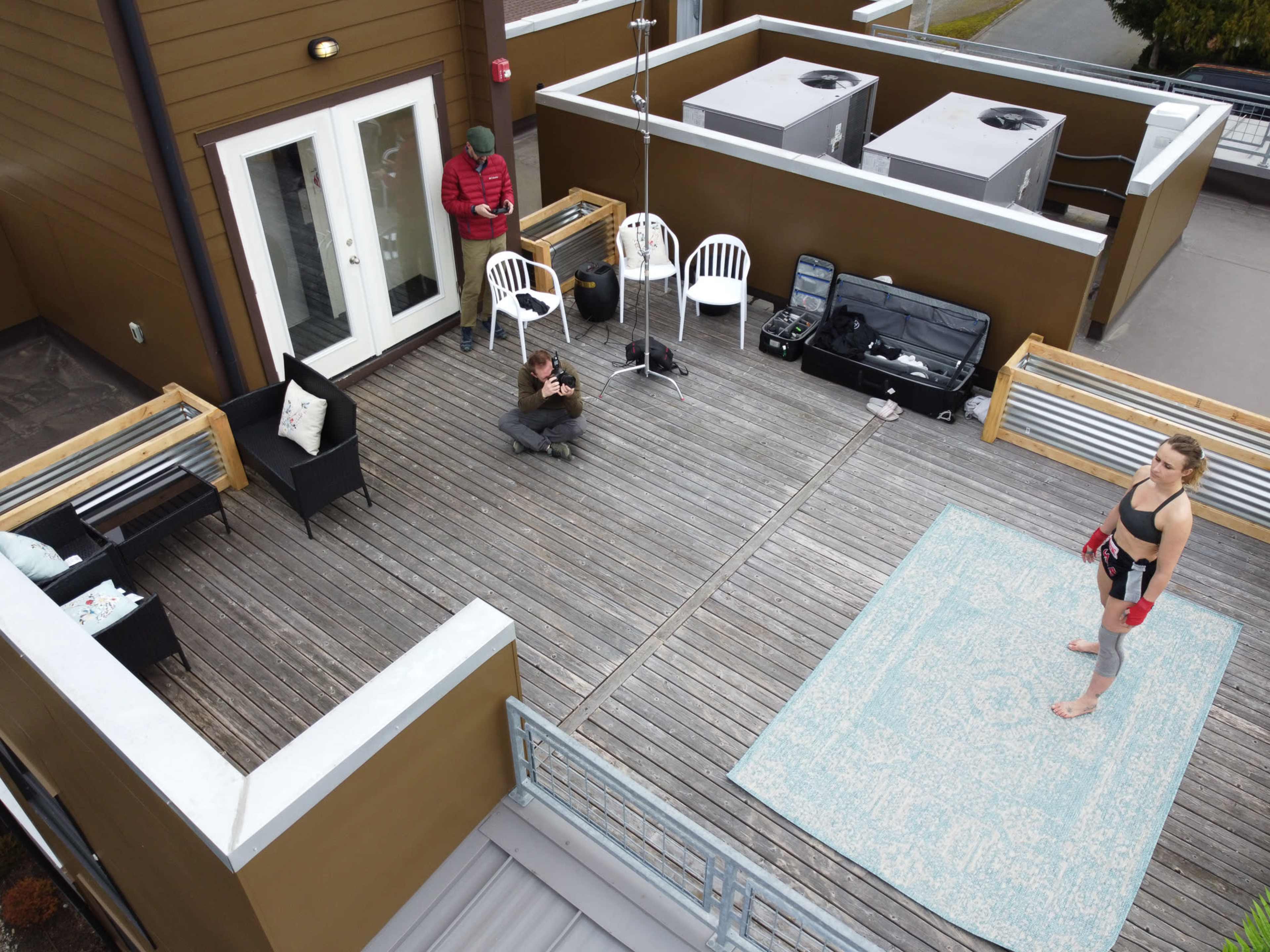 A wooden deck on a rooftop features a woman in workout attire standing on a rug, while two other individuals are positioned in different areas of the space.