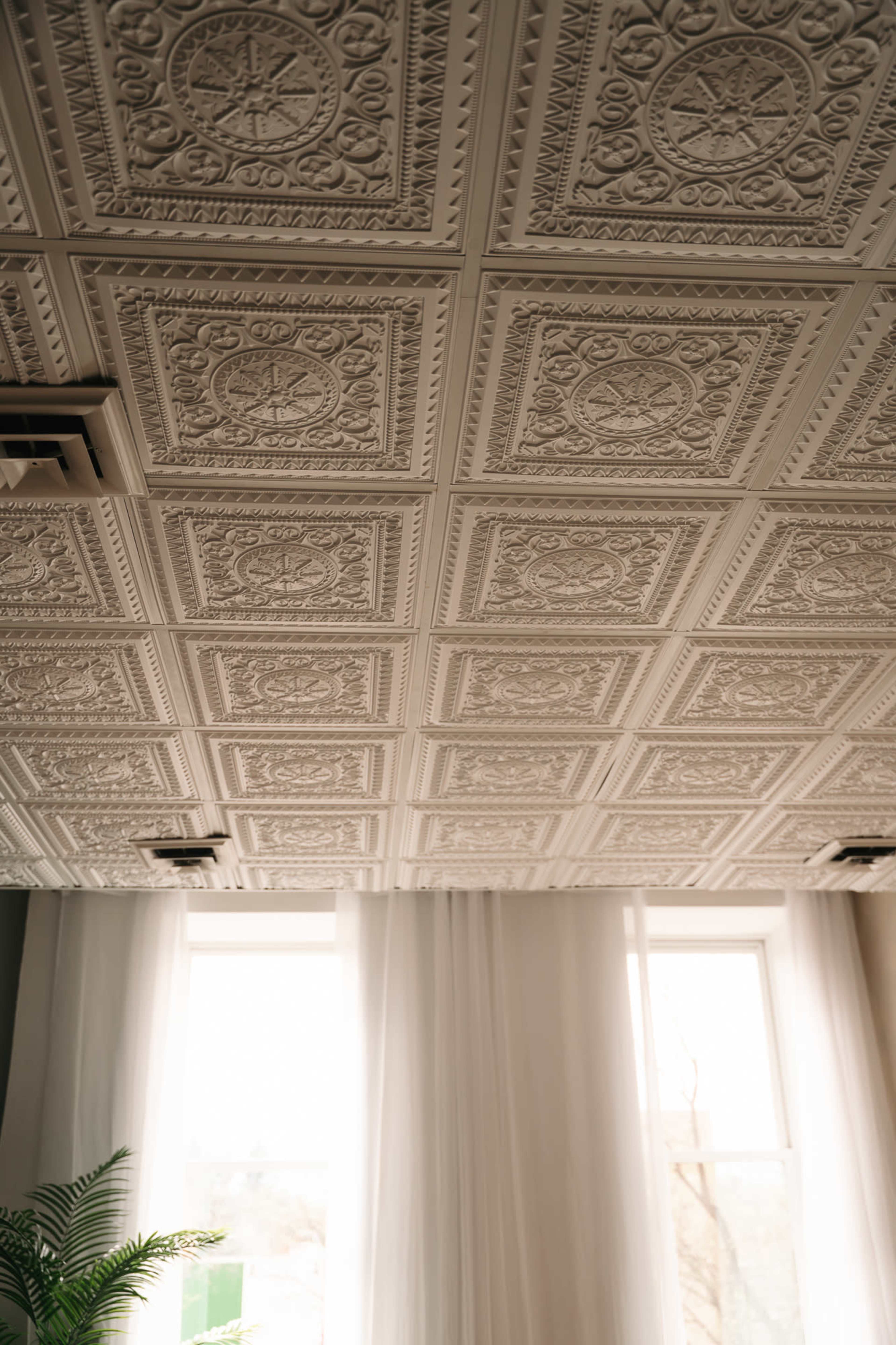 The image shows a close-up view of an elaborately textured ceiling with decorative panels and large windows that allow natural light to enter the room.