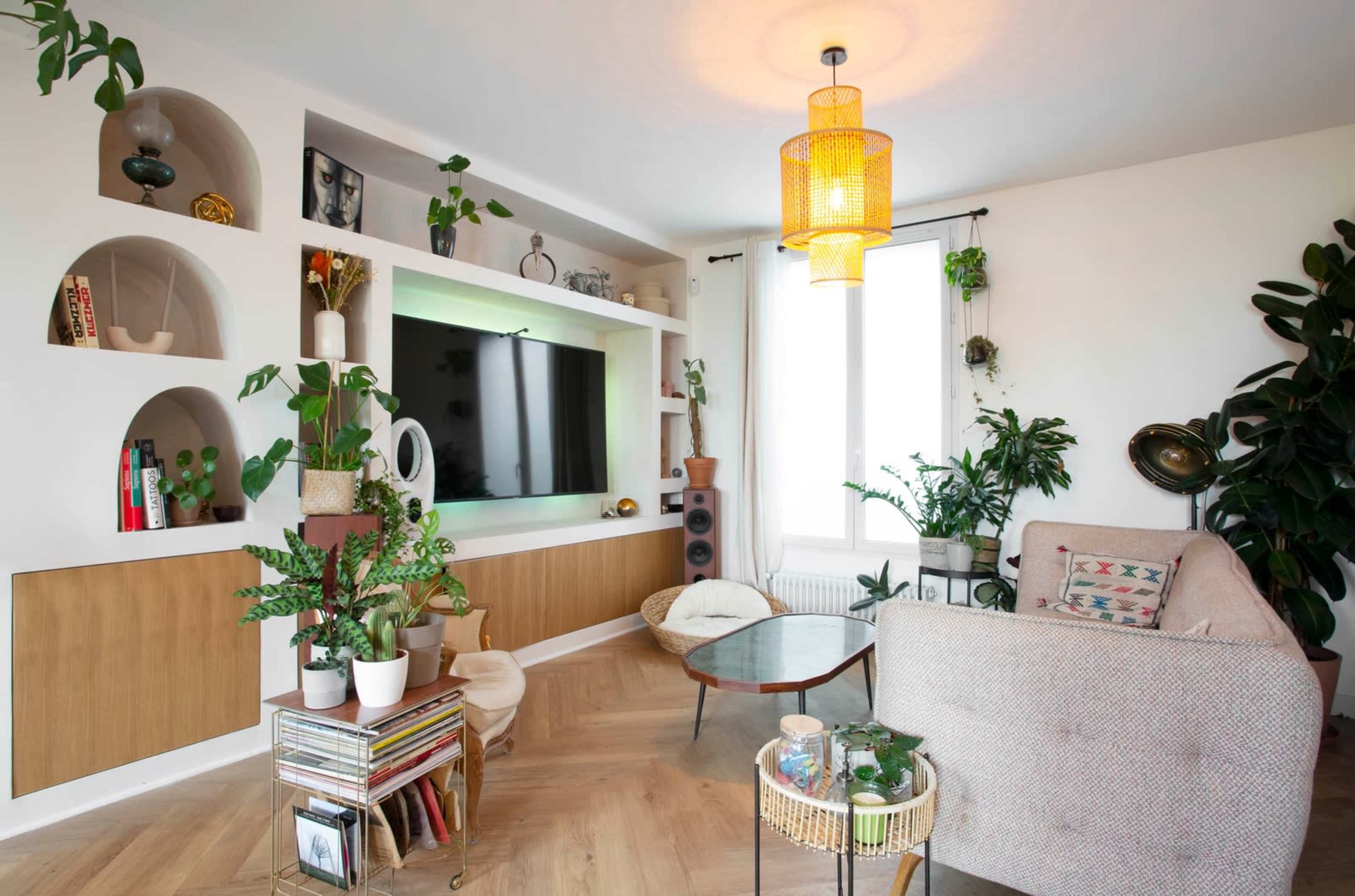 A modern living room features a wall-mounted television, shelves filled with decor, and various plants alongside a cozy seating area and a circular coffee table.