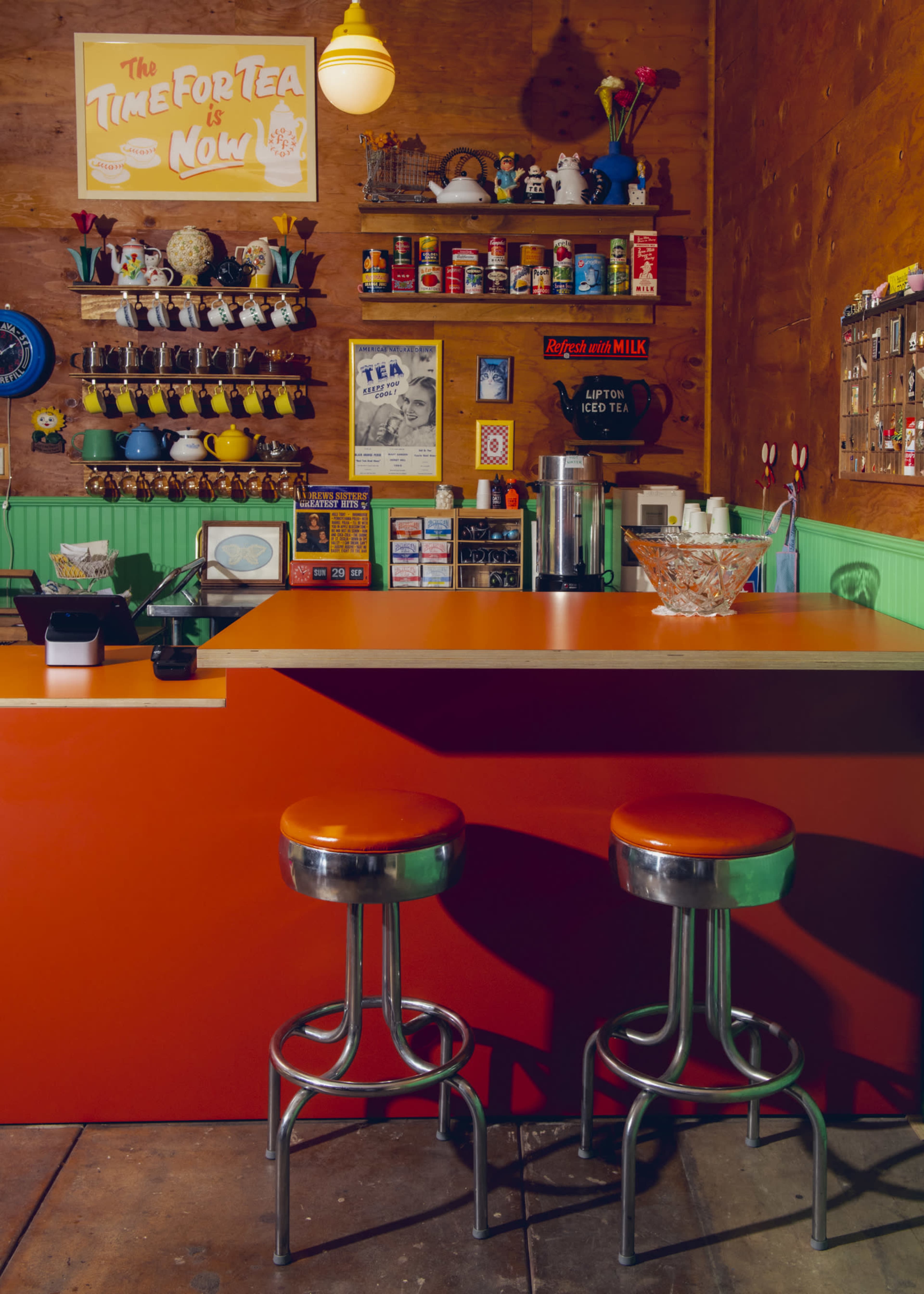 The image shows a brightly colored tea shop interior with an orange countertop, metal bar stools, and shelves lined with various tea-related items and decorations.