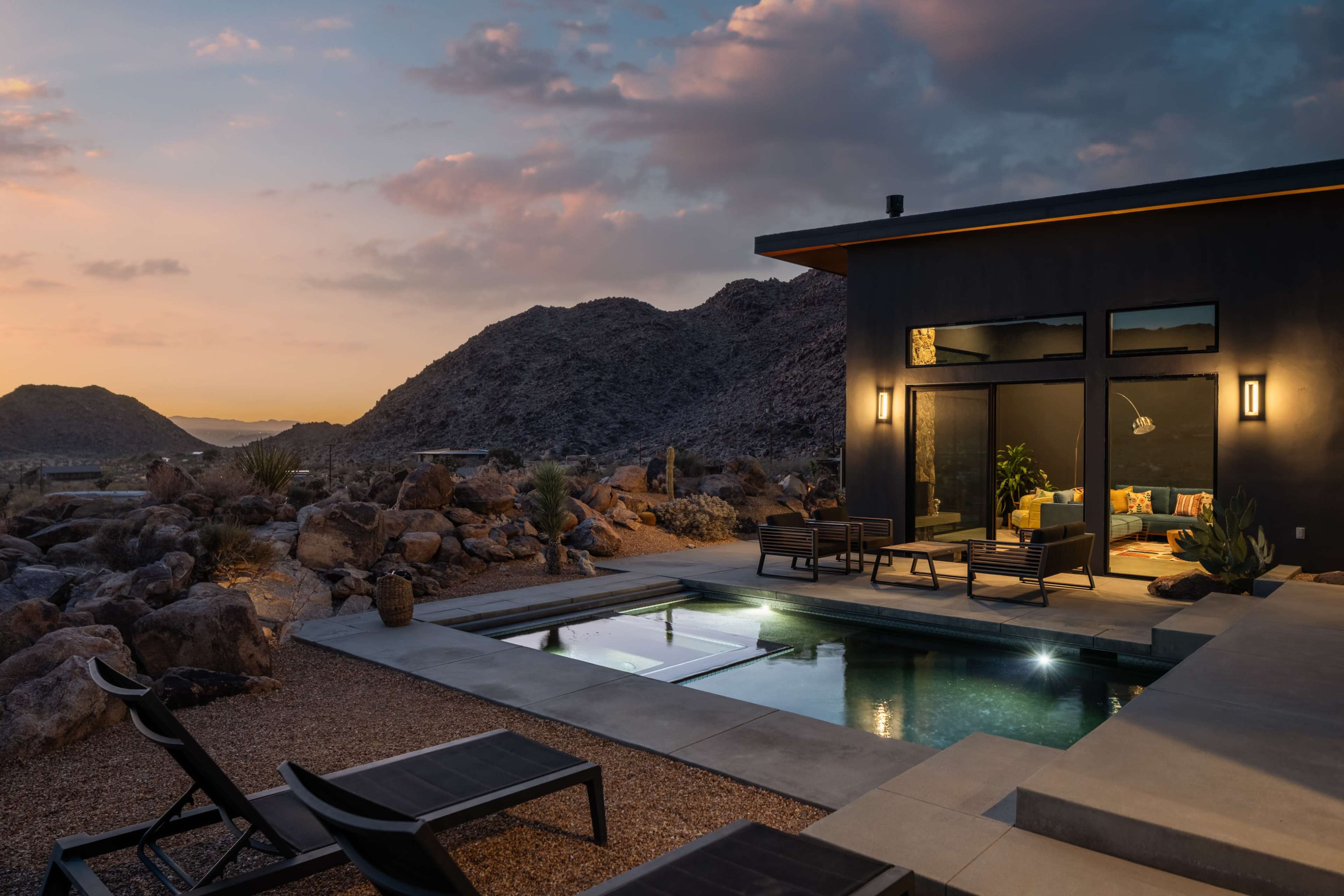The image shows a modern house with a pool surrounded by rocky terrain and desert hills at dusk.