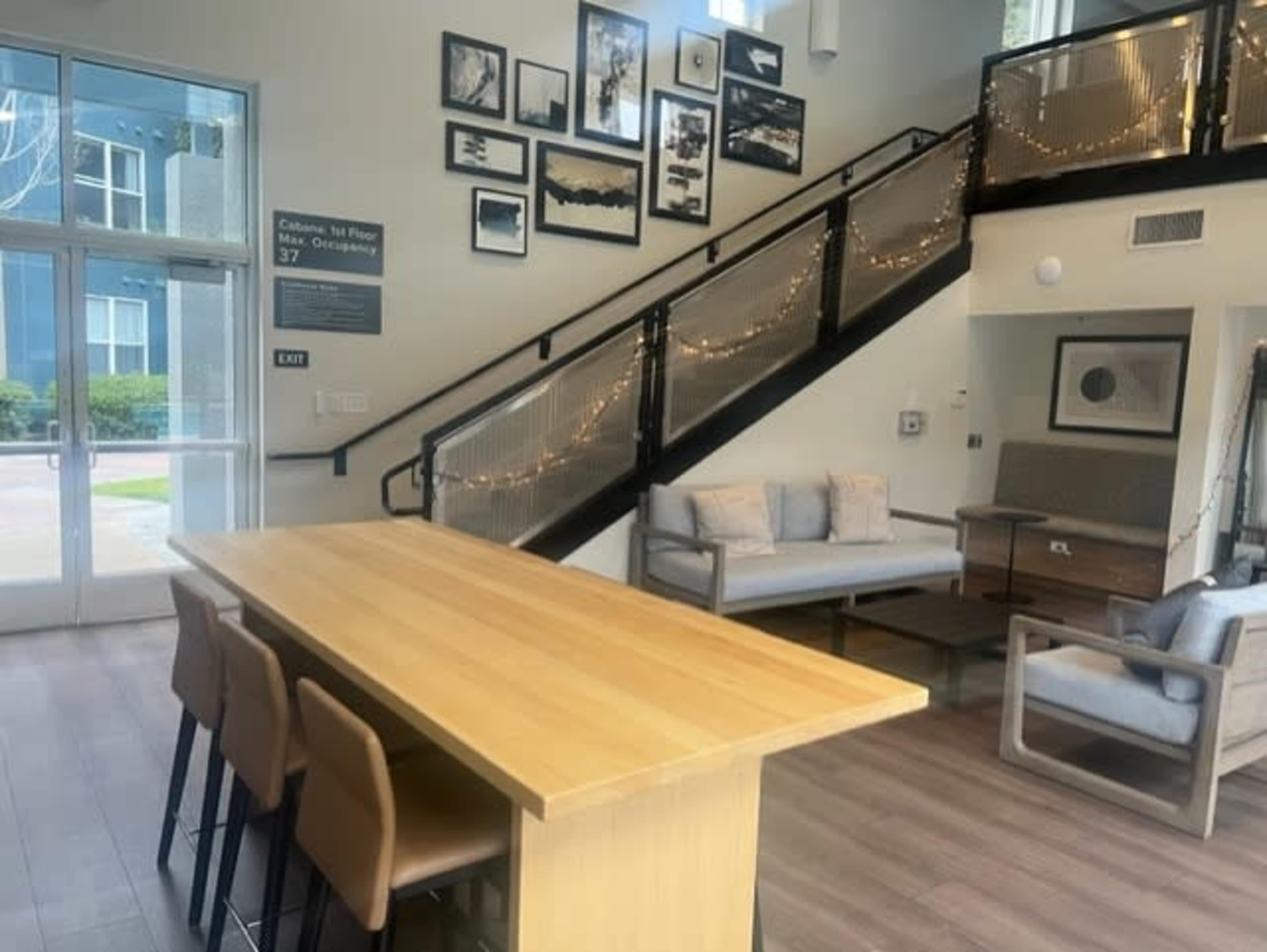 A modern indoor space featuring a wooden table with stools, a seating area with sofas, and a staircase adorned with framed pictures and decorative lighting.