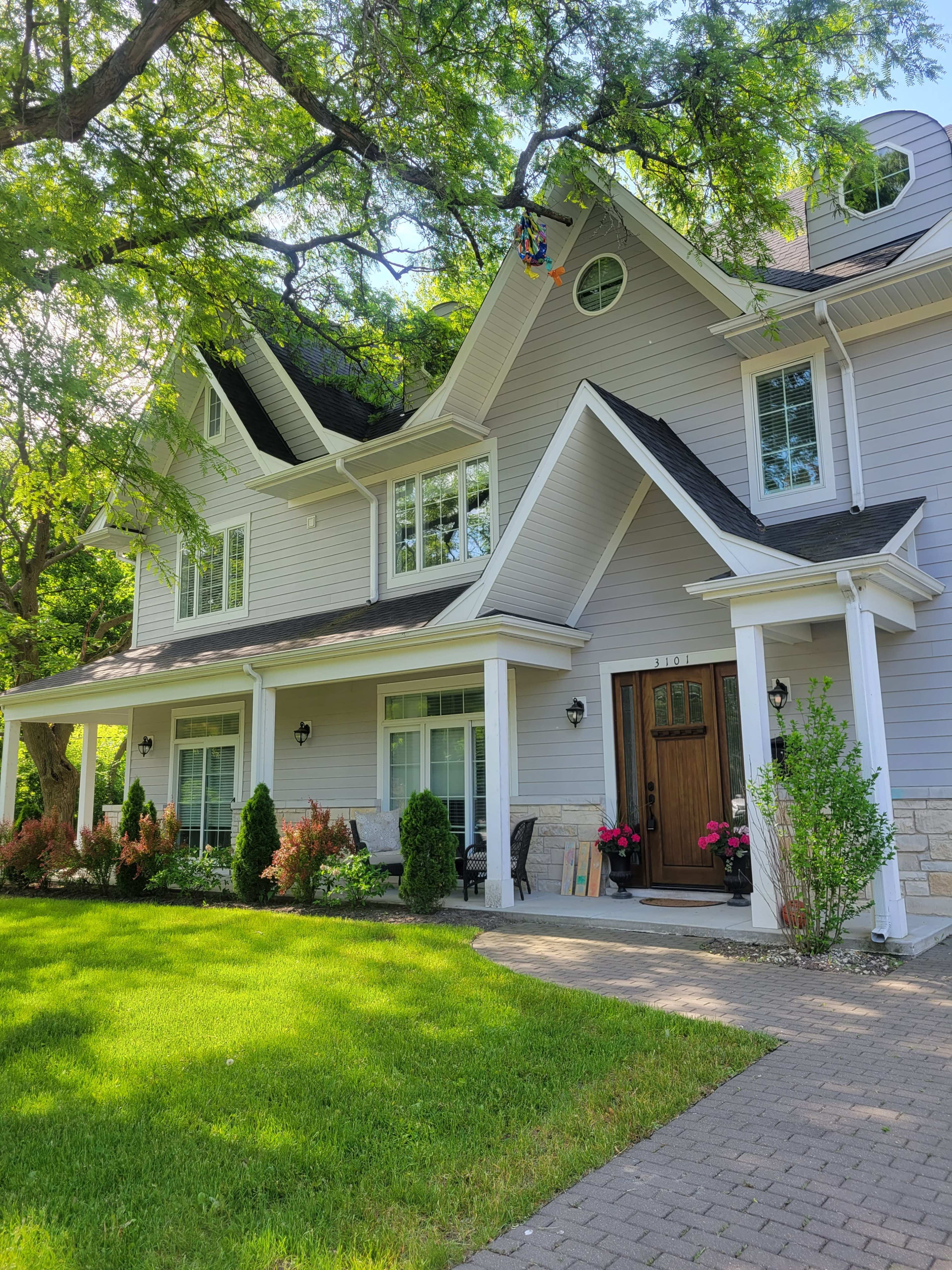 A large, two-story gray house with white trim features multiple windows and a wooden front door, surrounded by a well-maintained lawn and landscaped garden.